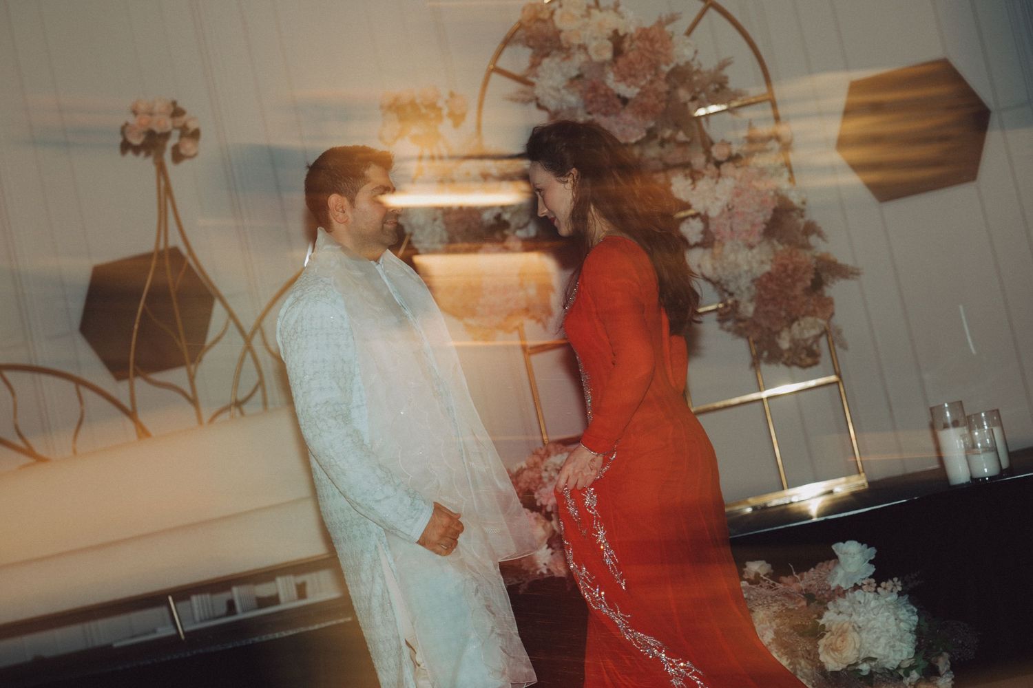 Couple in traditional South Asian attire, man in light sherwani and woman in red lehenga, at an elegant event.
