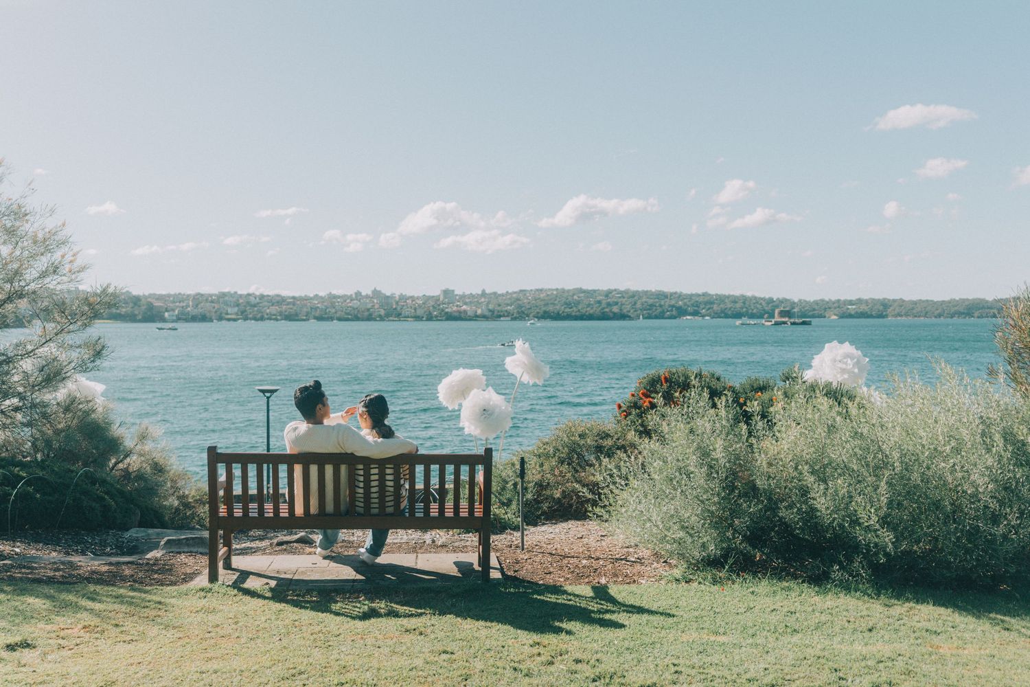 Couple sitting on a wooden bench overlooking a serene lake surrounded by lush green trees on a sunny day.