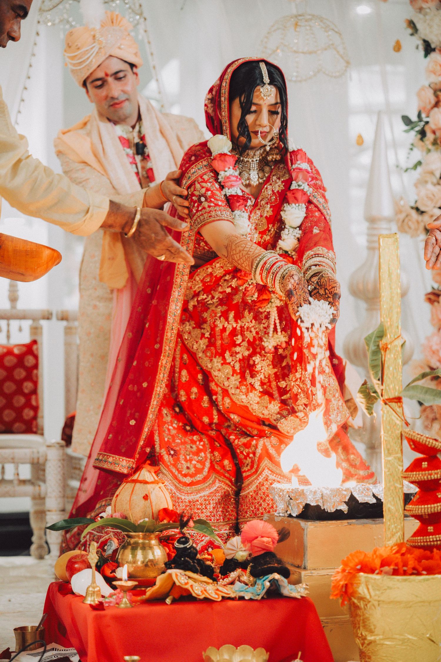 Bride in red lehenga and groom in cream sherwani performing Hindu wedding rituals around sacred fire.