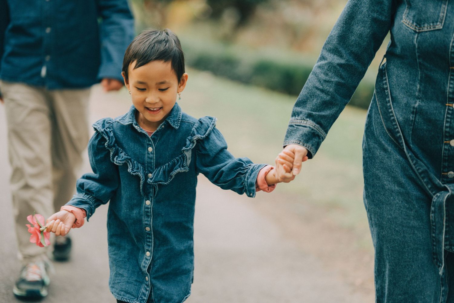 A child in denim outfit holds hands with adults while walking outdoors on a path.