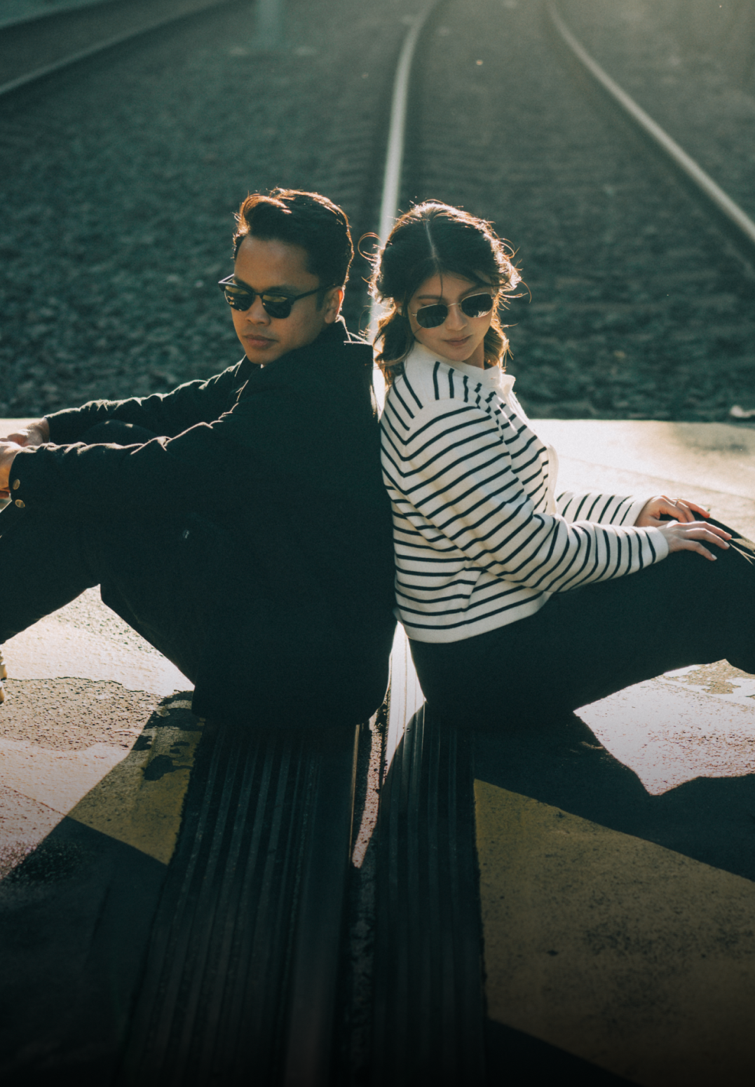Two people in striped shirts and sunglasses posing dramatically on train tracks at sunset.