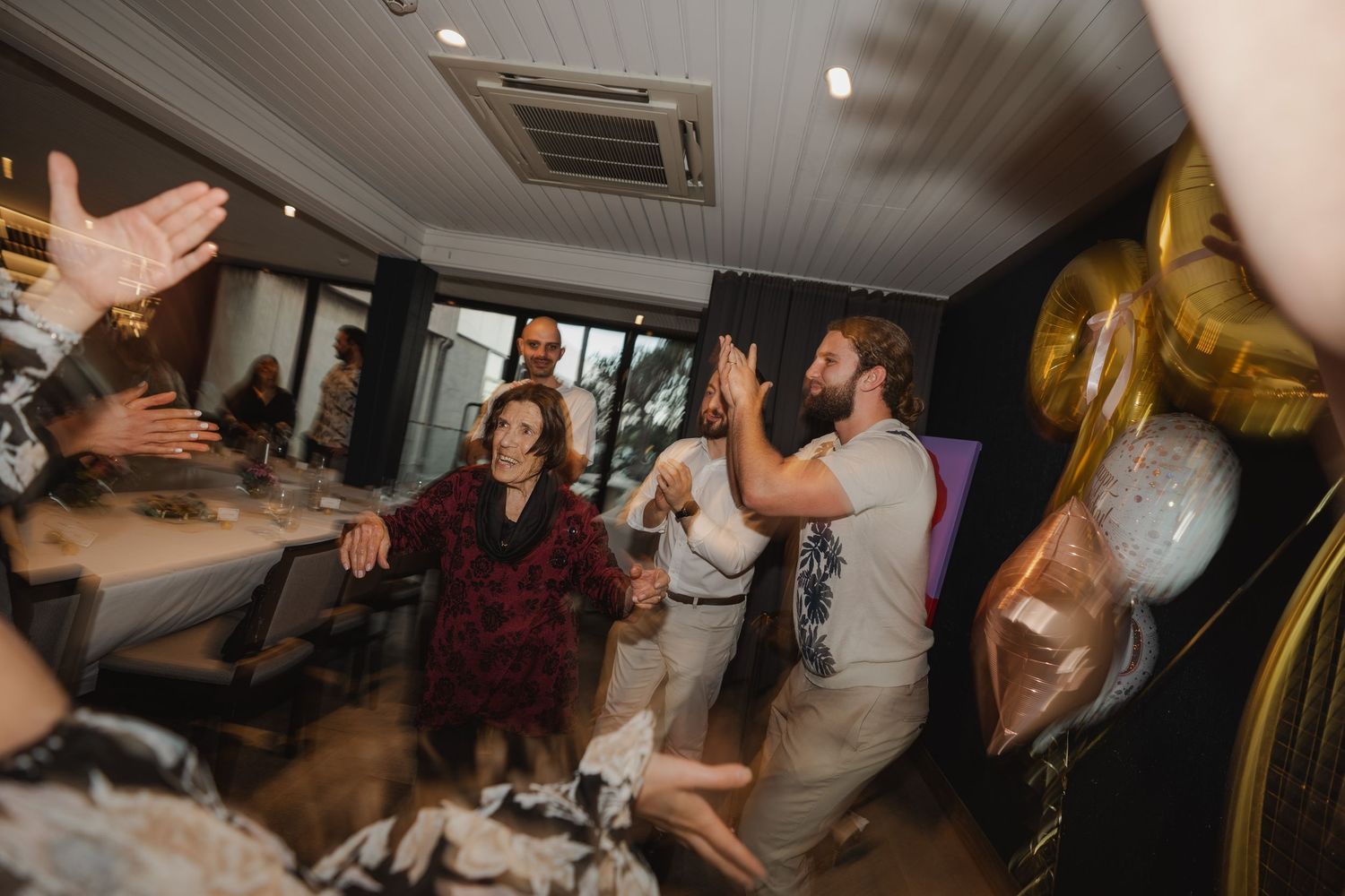 People dancing and celebrating at an indoor party with gold balloons visible in the background.