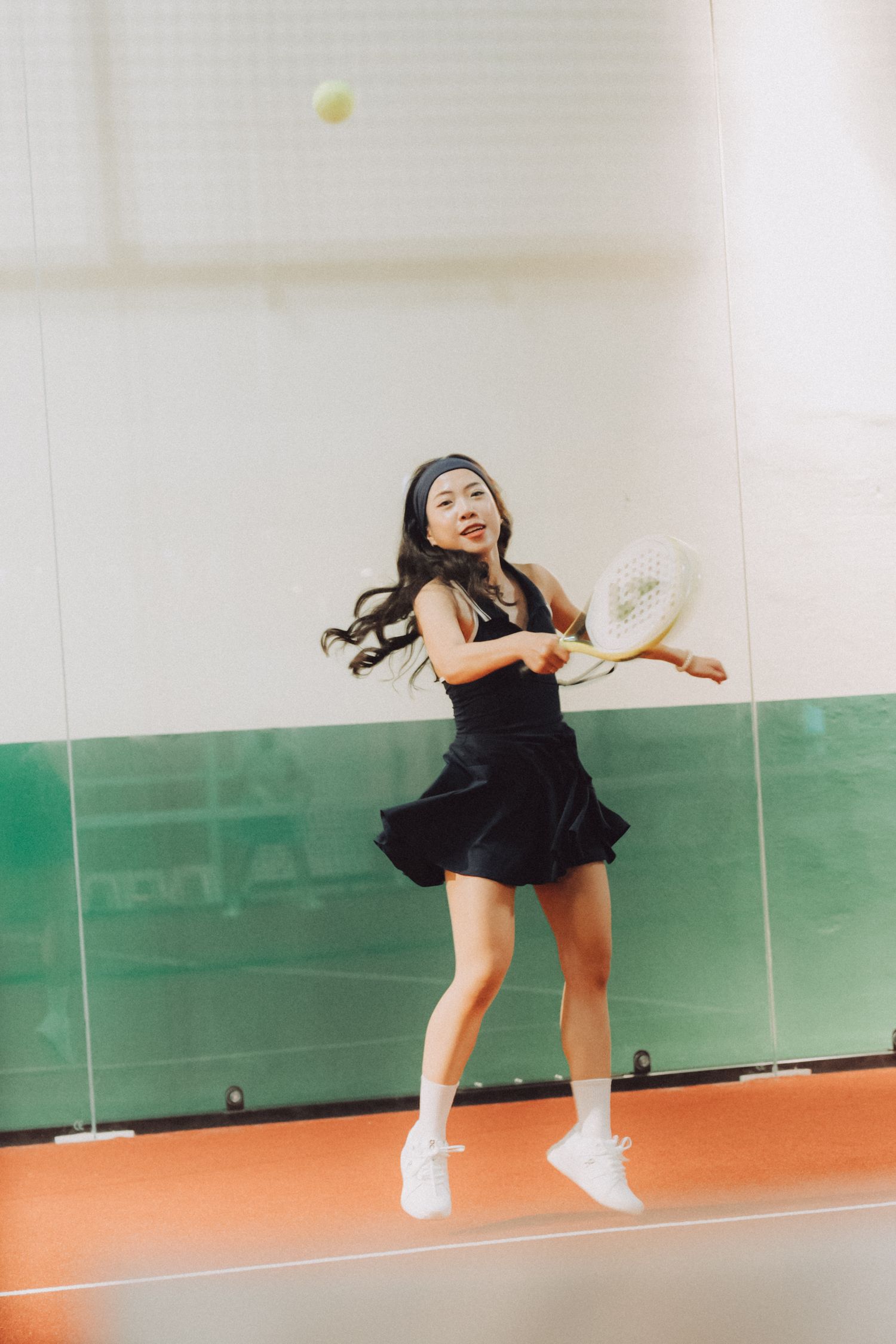 A person in a black dress and white shoes performs a dynamic jumping motion sequence on an indoor tennis court.