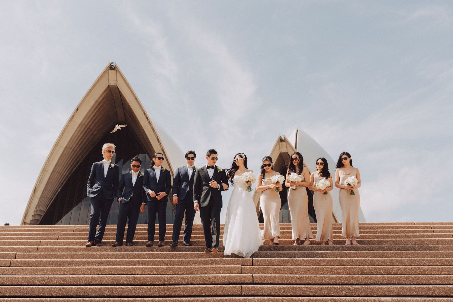 Wedding party members pose on grand stone steps outside a triangular architectural structure.