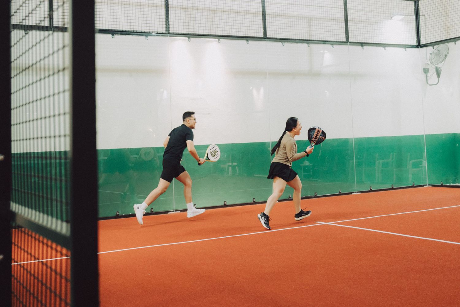 Two athletes perform agility drills on an indoor tennis court with green walls and red clay surface.