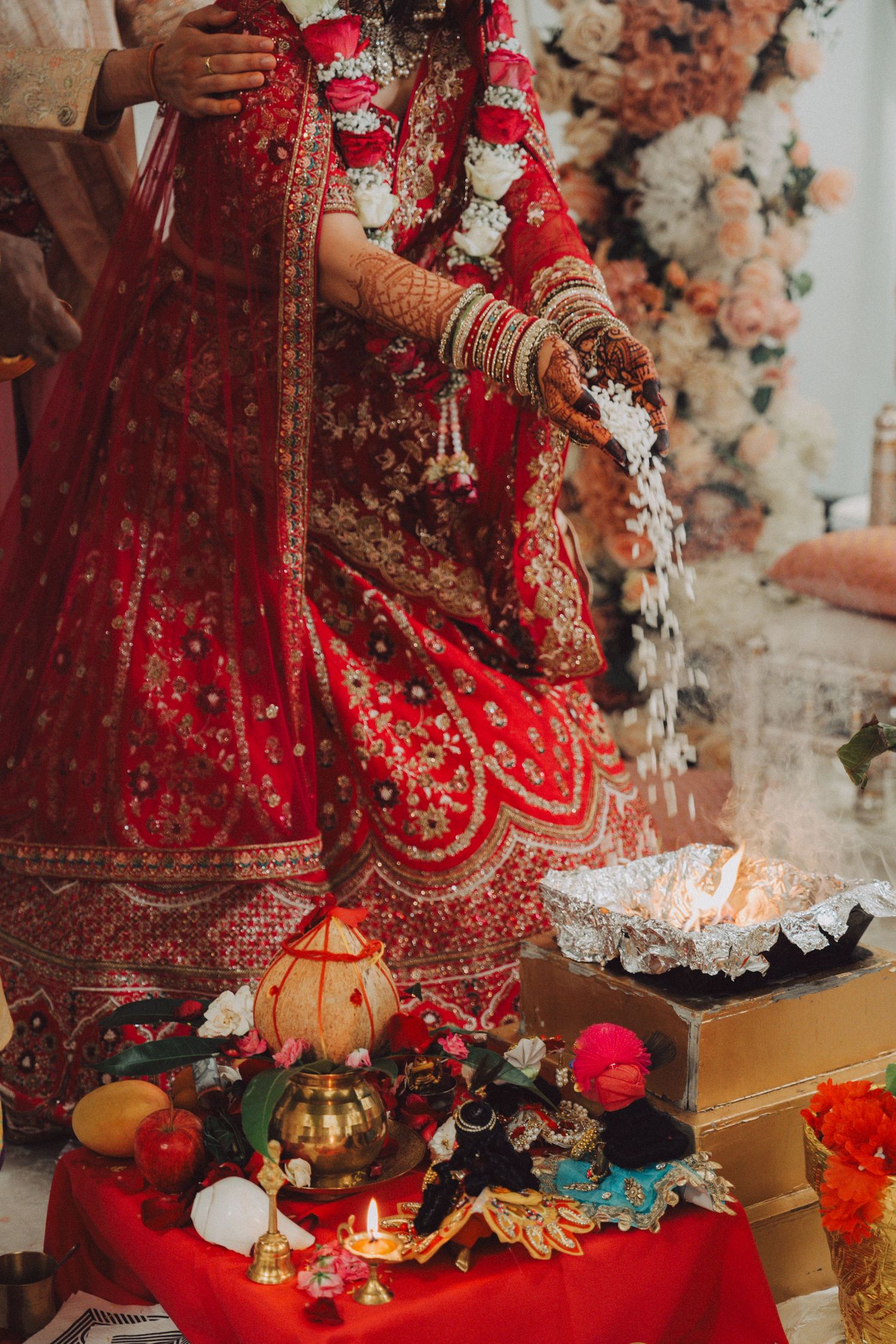 Bride in red lehenga performing Hindu wedding rituals by sacred fire with traditional offerings and floral decor.
