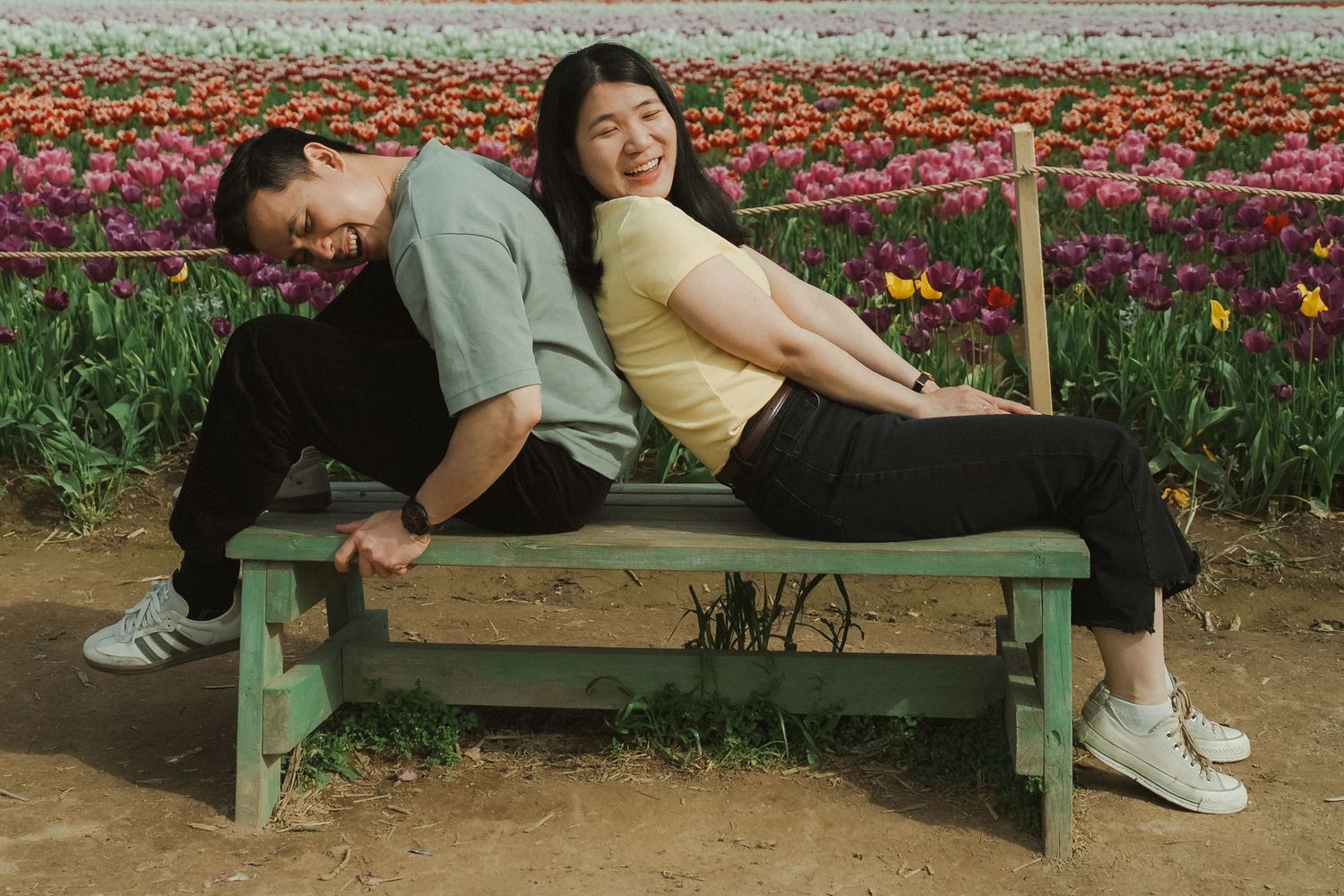 Two people sitting back-to-back on a green bench in a colorful tulip field, smiling and relaxed.