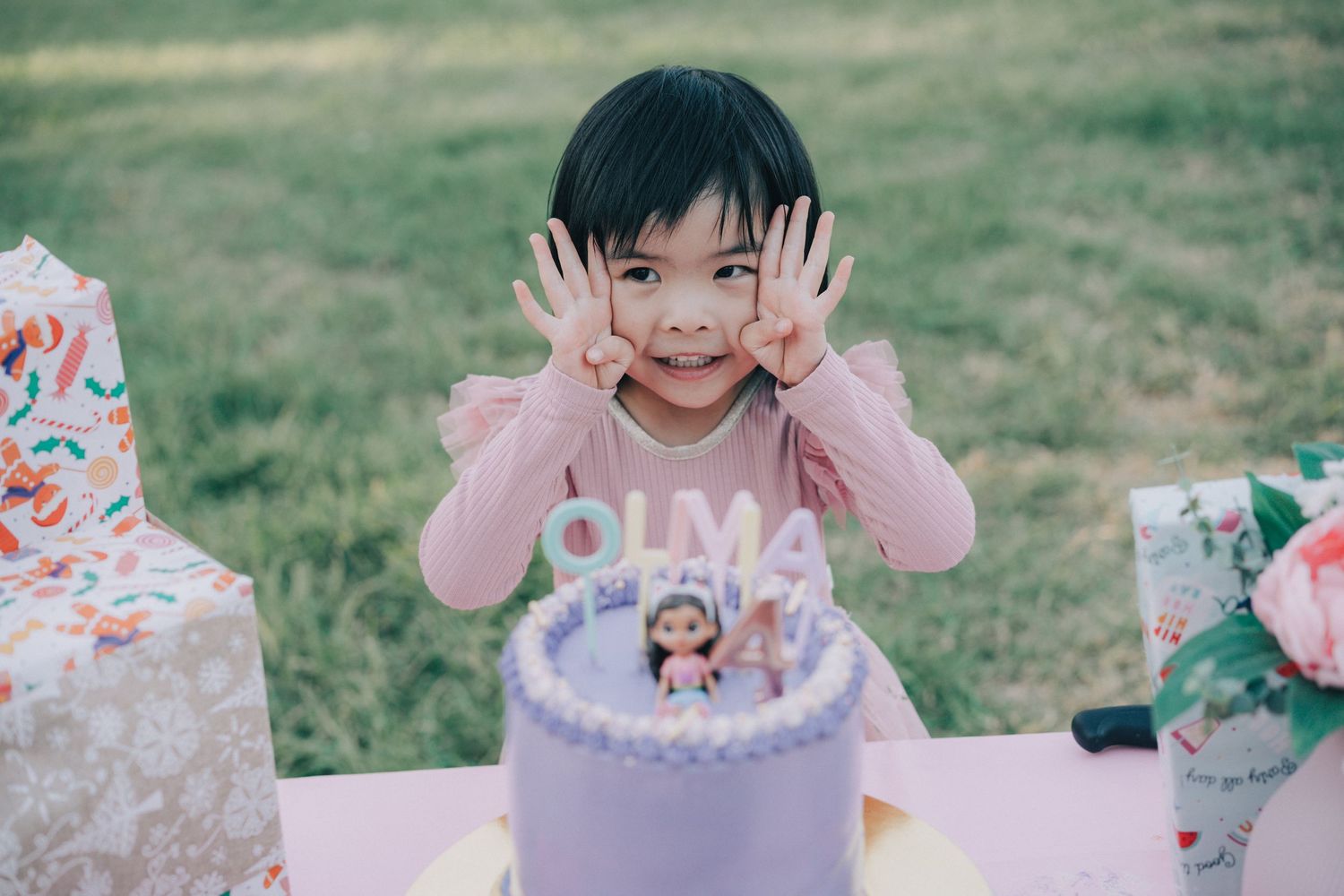 Children celebrate at an outdoor birthday party with pink decorations and a festive cake on a sunny day.