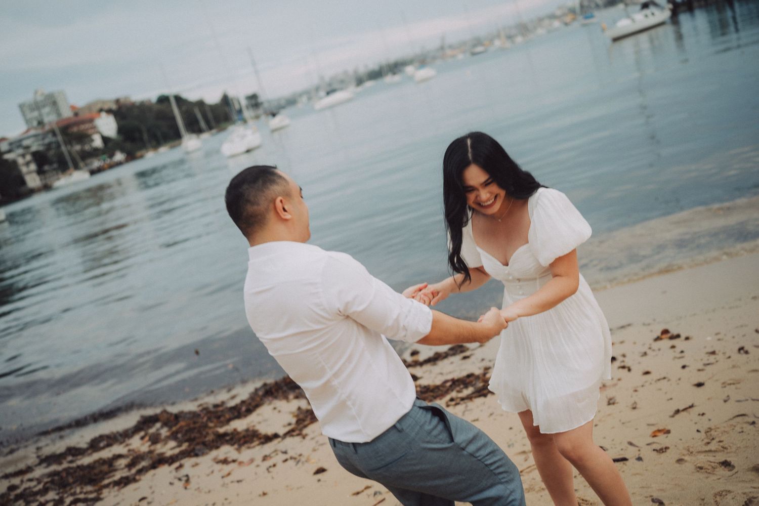 A couple in white outfits shares a playful moment on a beach with a city waterfront in the background.