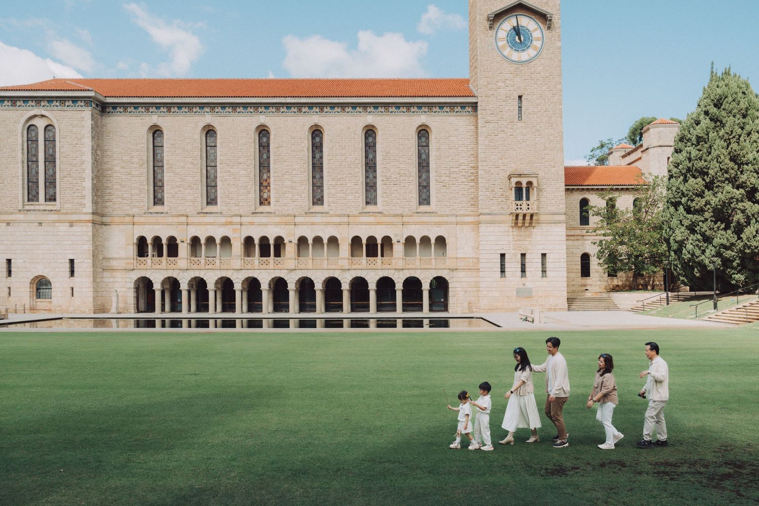 Players in white uniforms practice cricket on a lush green field next to a historic stone building with arched windows.