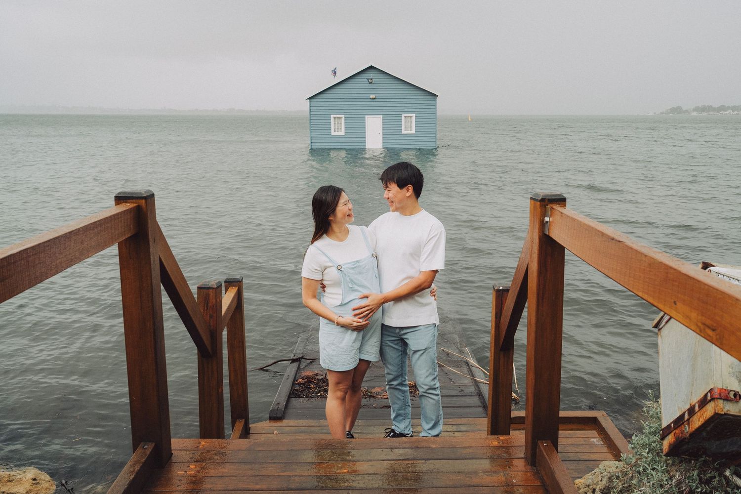 A couple sits together on a wooden pier overlooking the ocean with a blue house in the background.
