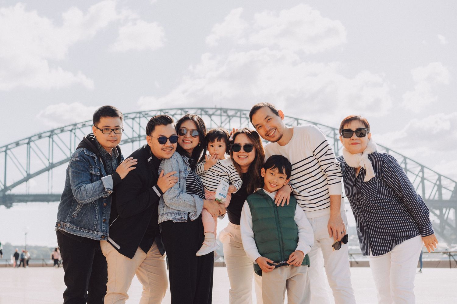 A group of people walking and posing together along a waterfront with a bridge visible in the background.