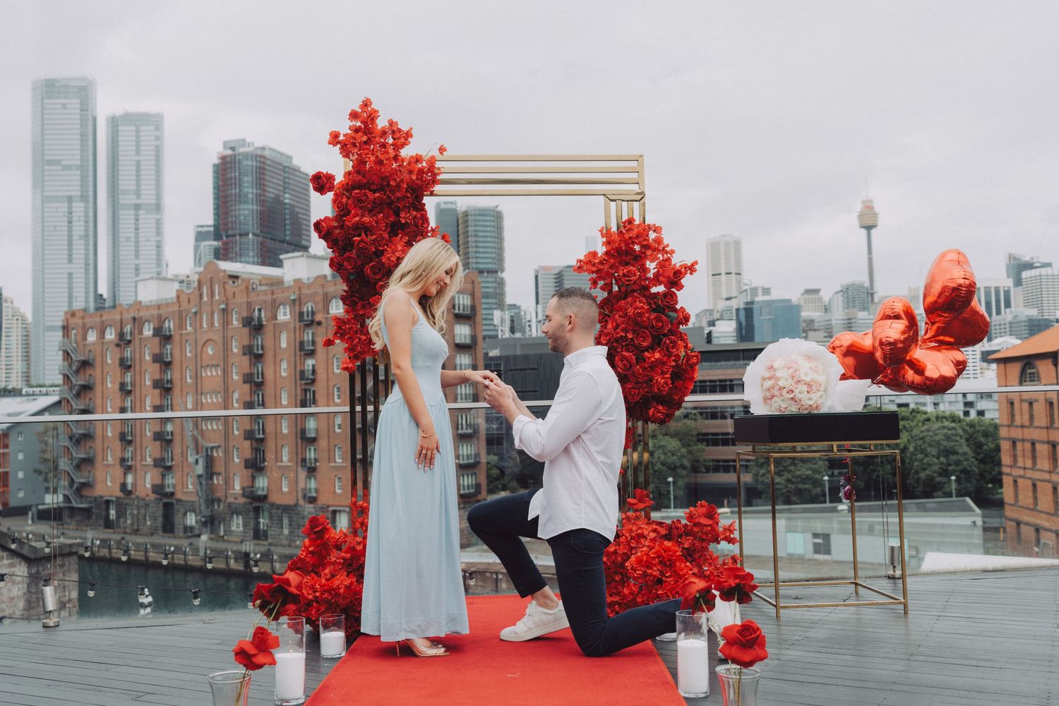 A romantic proposal scene with red heart balloons against a cityscape backdrop at sunset.