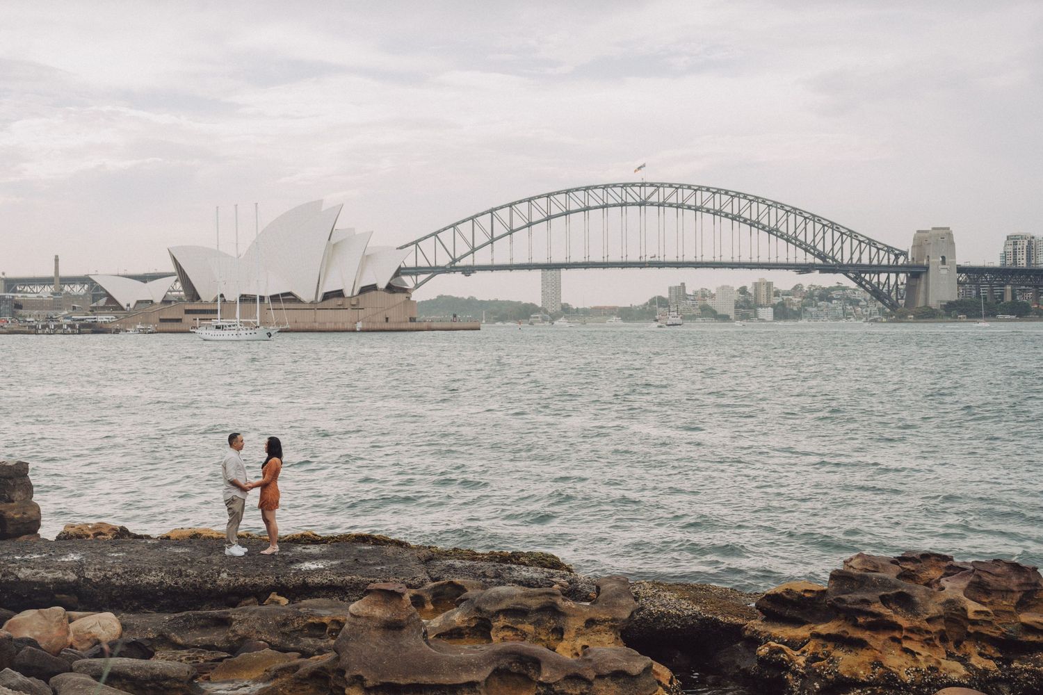 Scenic views of Sydney Harbour Bridge on a cloudy day with boats in the water and rocky shoreline in the foreground.