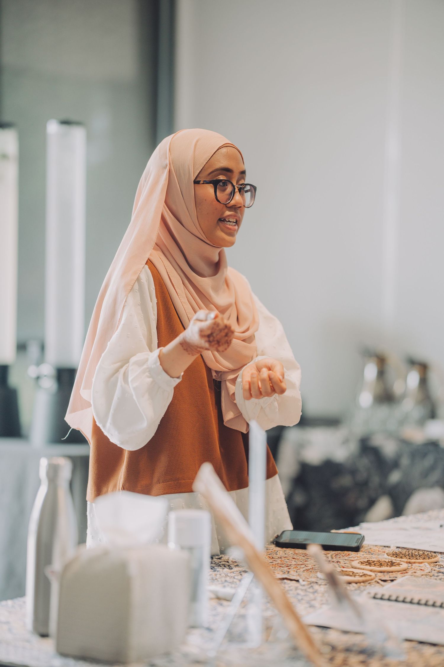 Muslim woman in hijab and glasses sits at desk with notebooks and study materials in bright modern workspace.