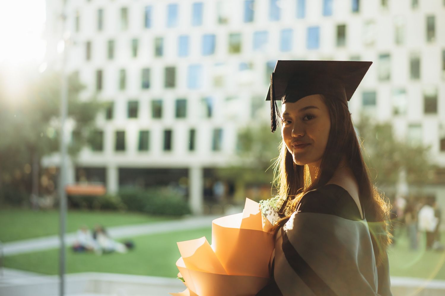 A graduate in black cap and gown holds a bouquet of orange flowers while standing outdoors.