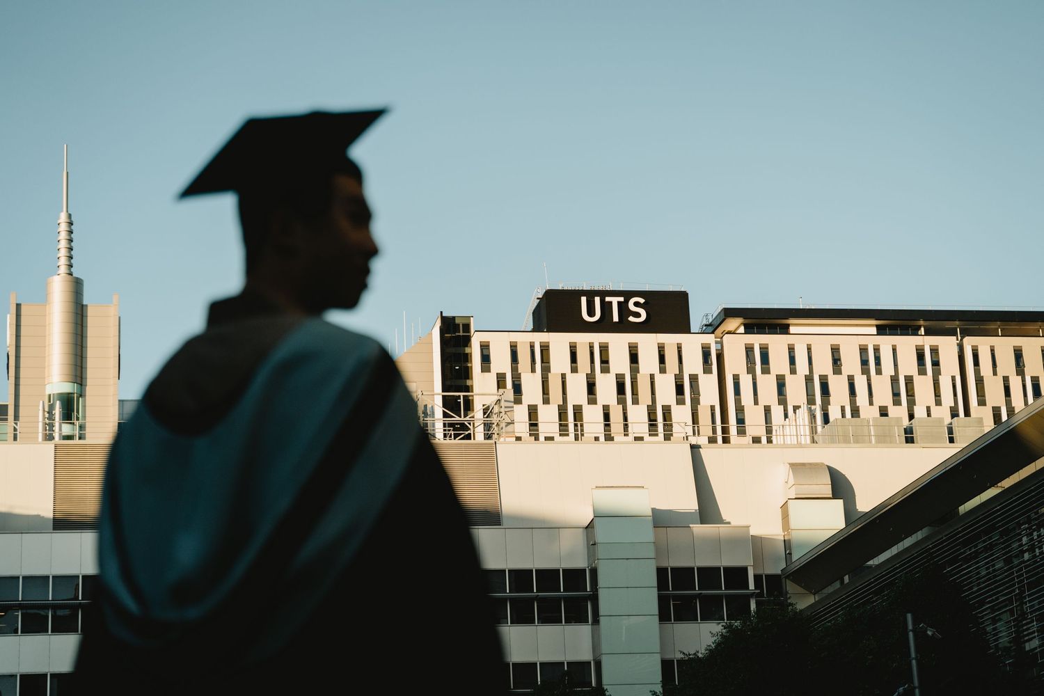 Graduates in blue caps and gowns stand against a backdrop of modern university buildings during commencement ceremony.