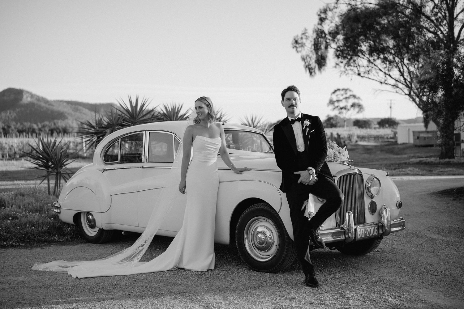 Classic vintage car serves as backdrop for romantic black and white wedding photography session in outdoor setting.