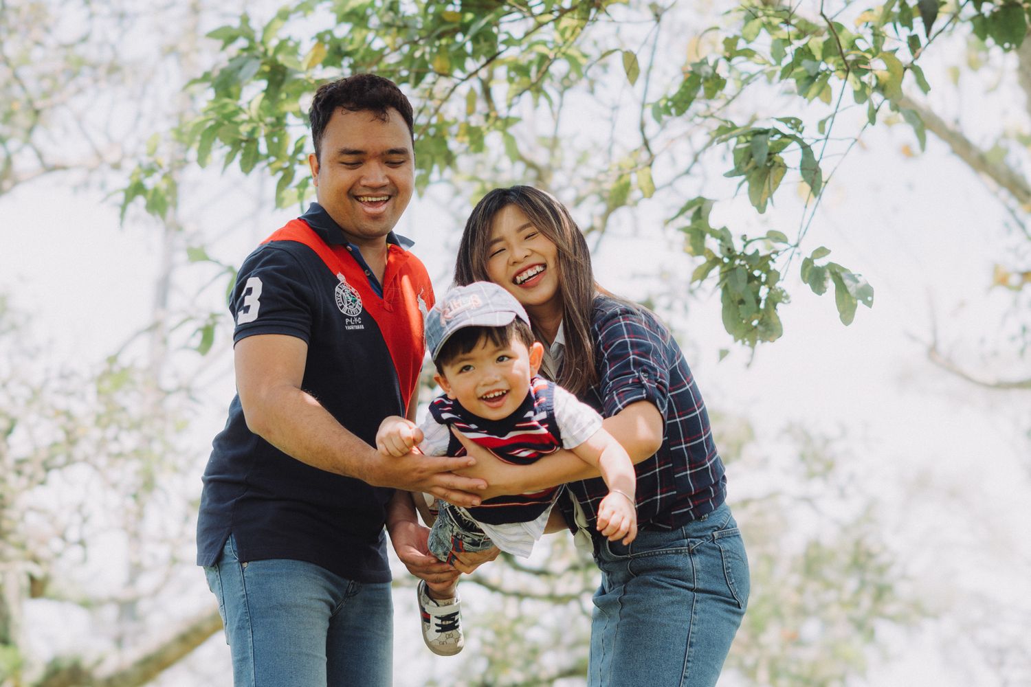 A family plays together outdoors on a bright day, lifting and swinging a small child while smiling and laughing.