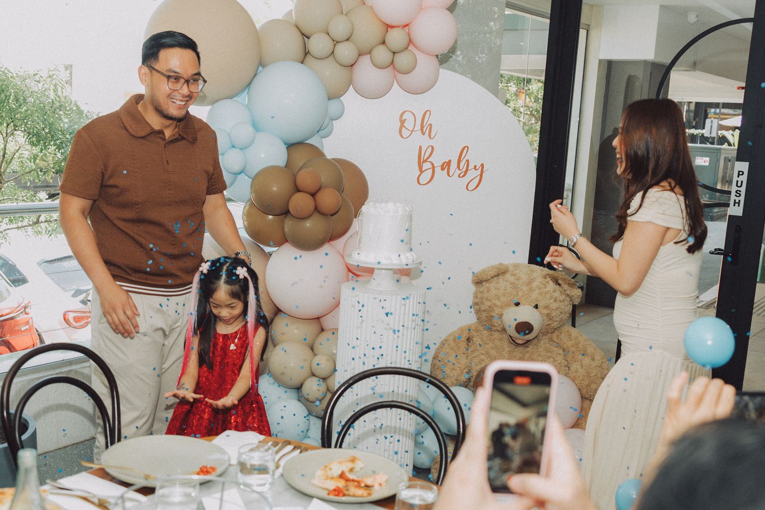 A baby shower celebration with balloon decorations, a teddy bear, and guests gathered around a decorated table.