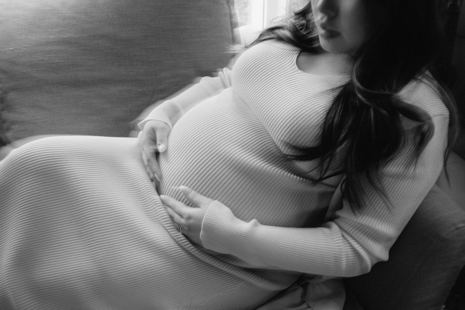 A black and white photograph of a pregnant woman in a light knit dress resting on a dark surface.