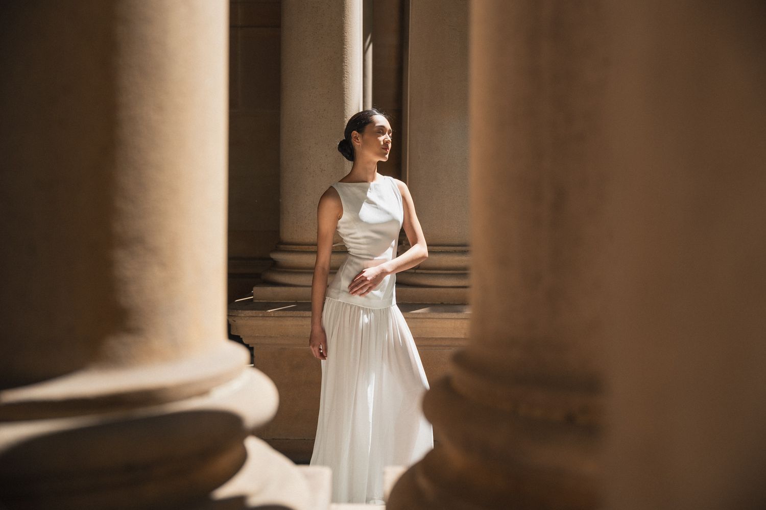 A bride in a white wedding gown stands elegantly between classical stone columns in dramatic lighting.