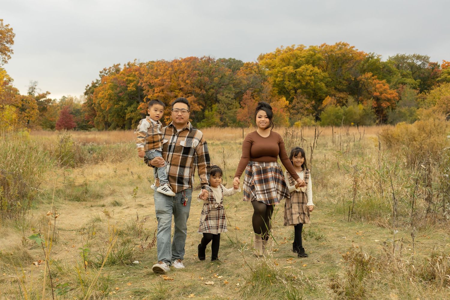 A group in coordinating plaid outfits walk together through a golden autumn field with trees in the background.