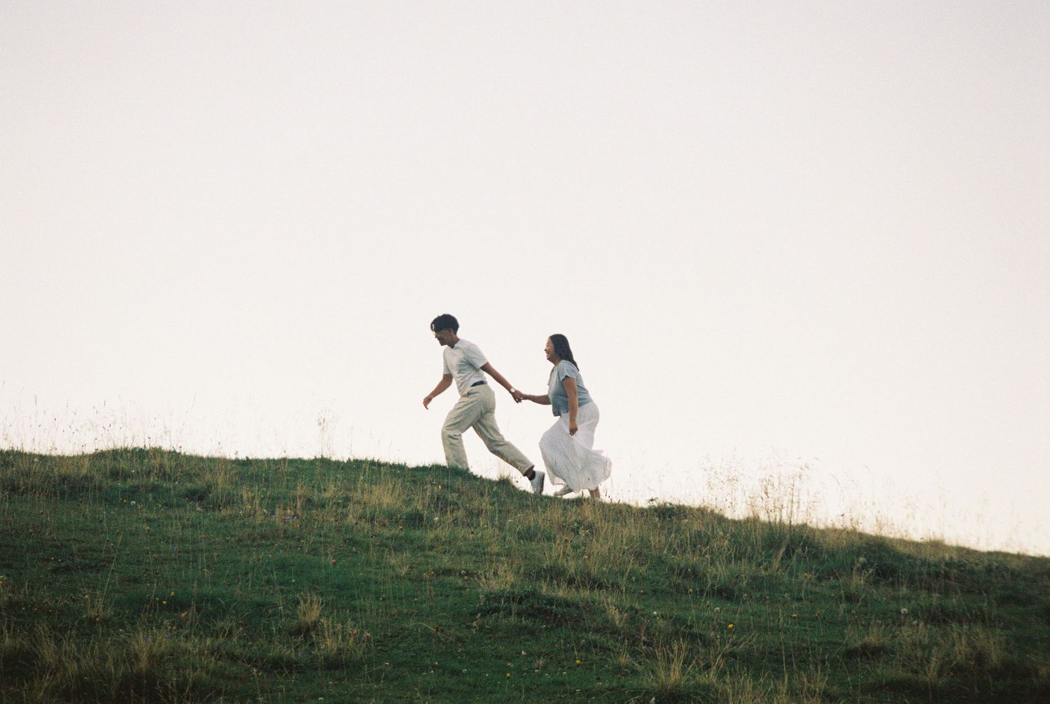 Two people run and play together on a grassy hillside against a bright sky at sunset.