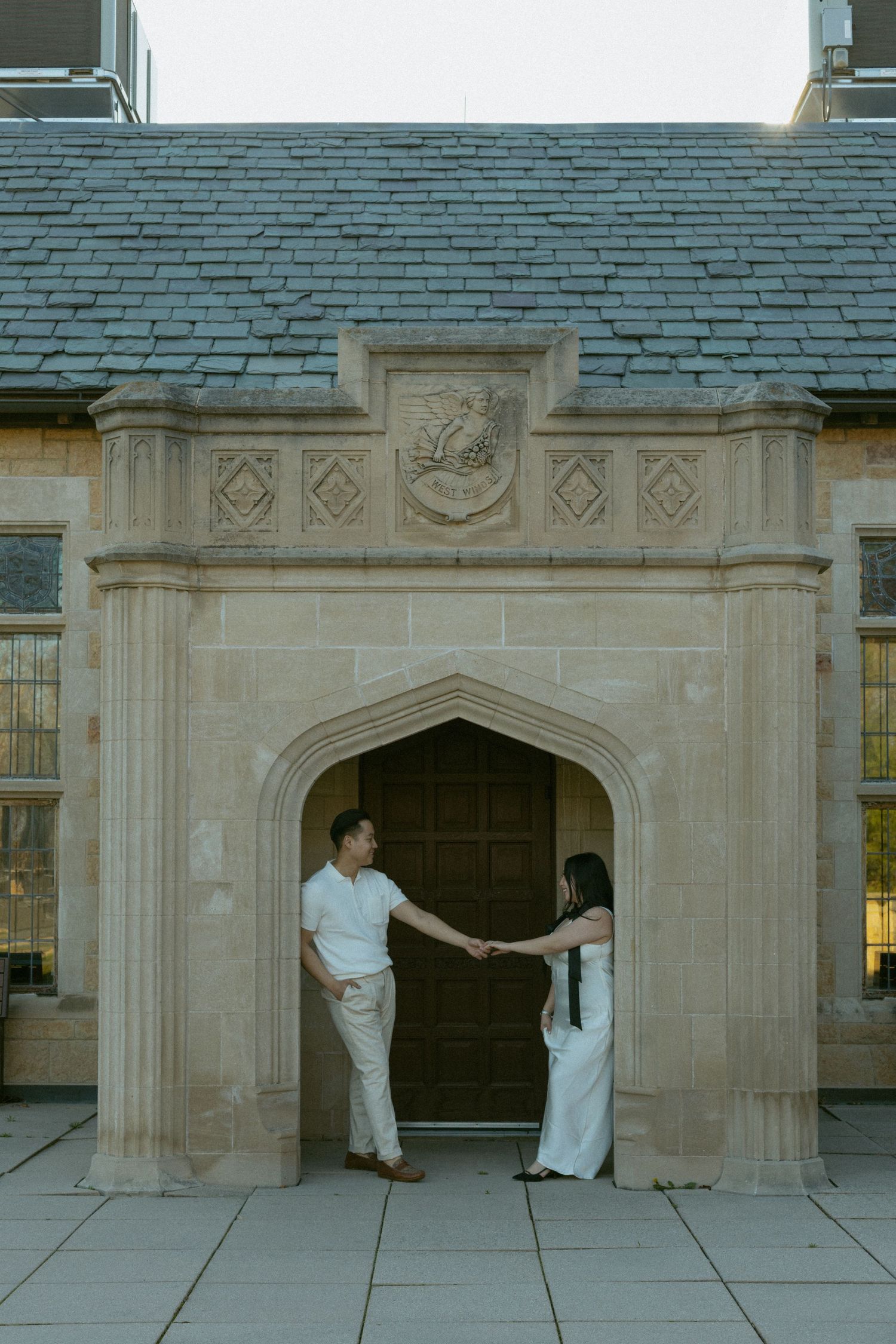 Two people in white clothing interact near an ornate stone archway with carved architectural details.