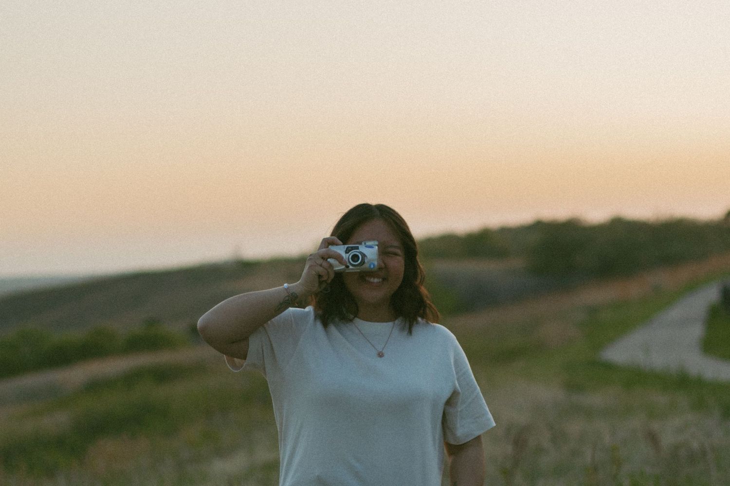 Person in sunglasses and white t-shirt standing in a field at sunset with winding road in background.
