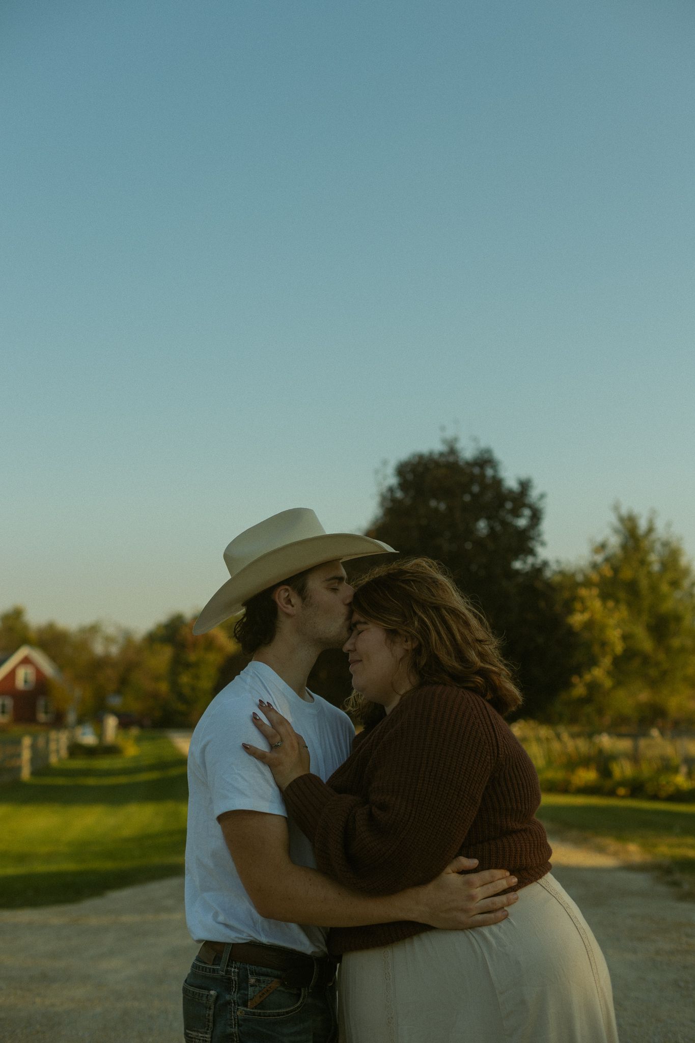 A romantic couple in cowboy attire embraces during sunset in a rural setting with trees and buildings in background.