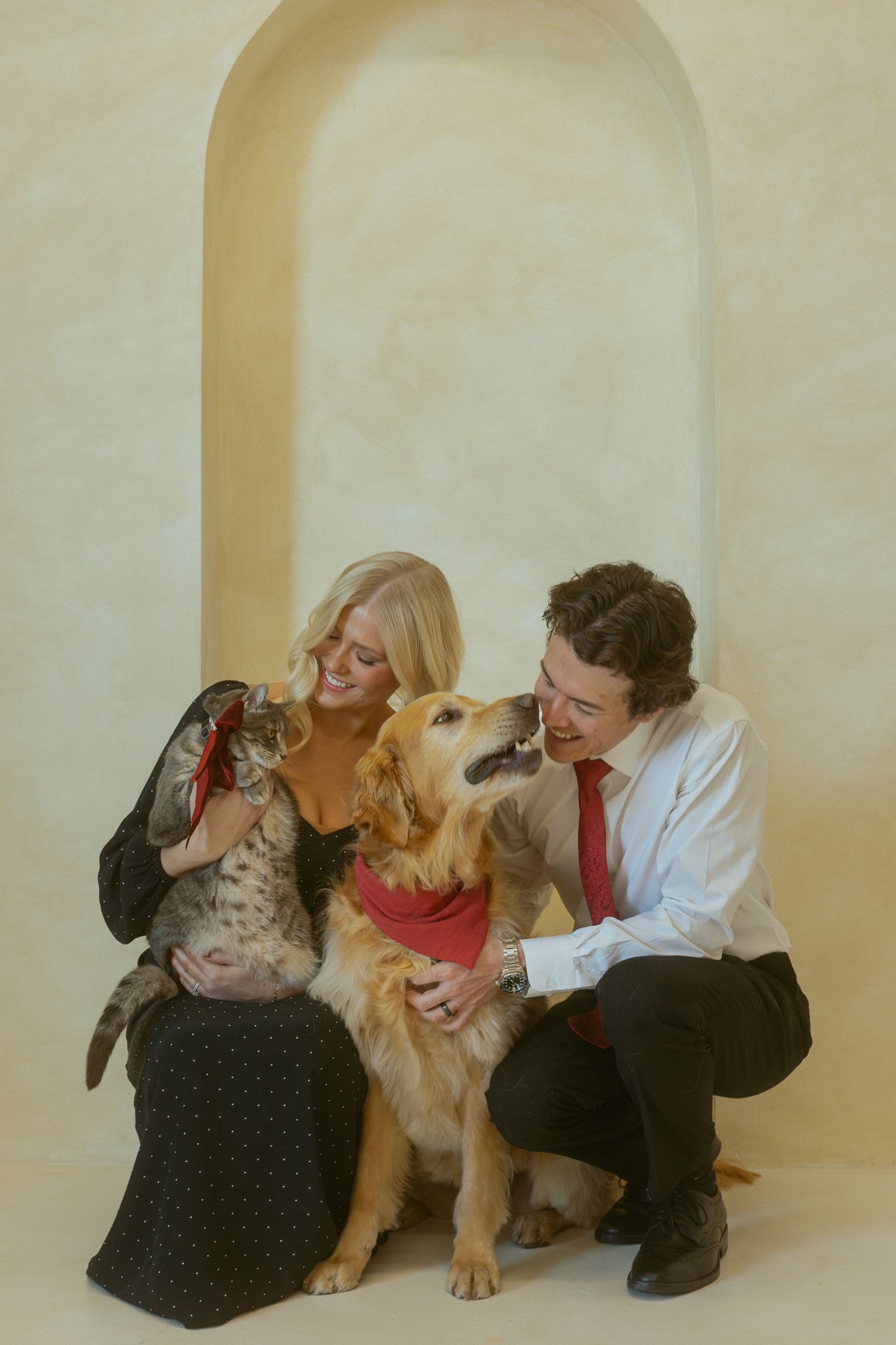 A golden retriever dog gets affectionate kisses and attention from two people in a studio setting with arched background.