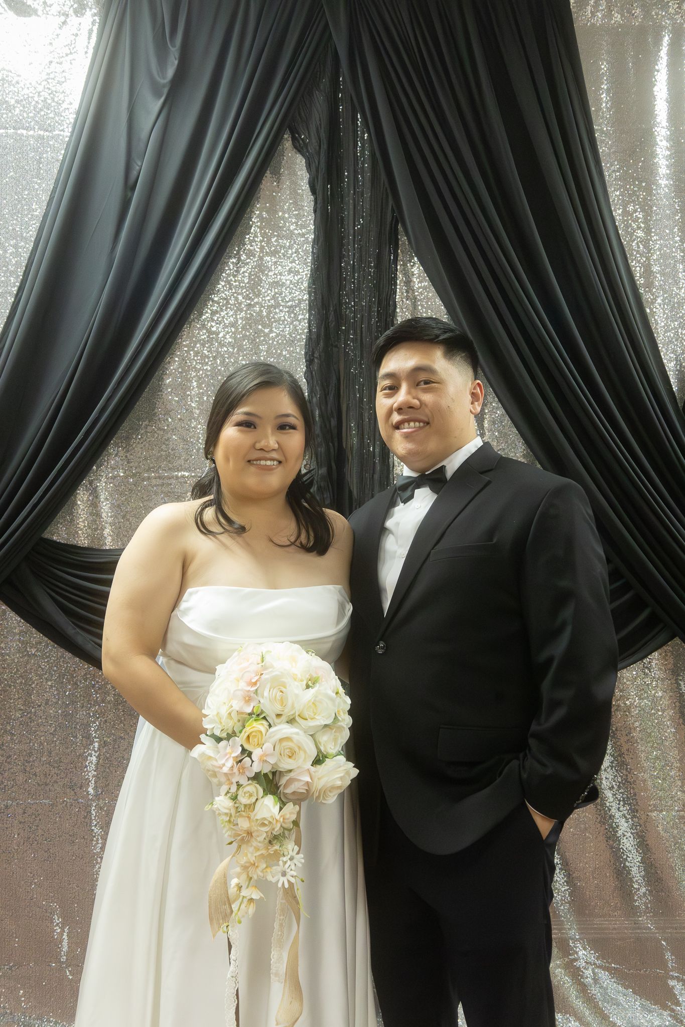 A couple in wedding attire share joyful moments against a backdrop of dark curtains during their wedding photoshoot.