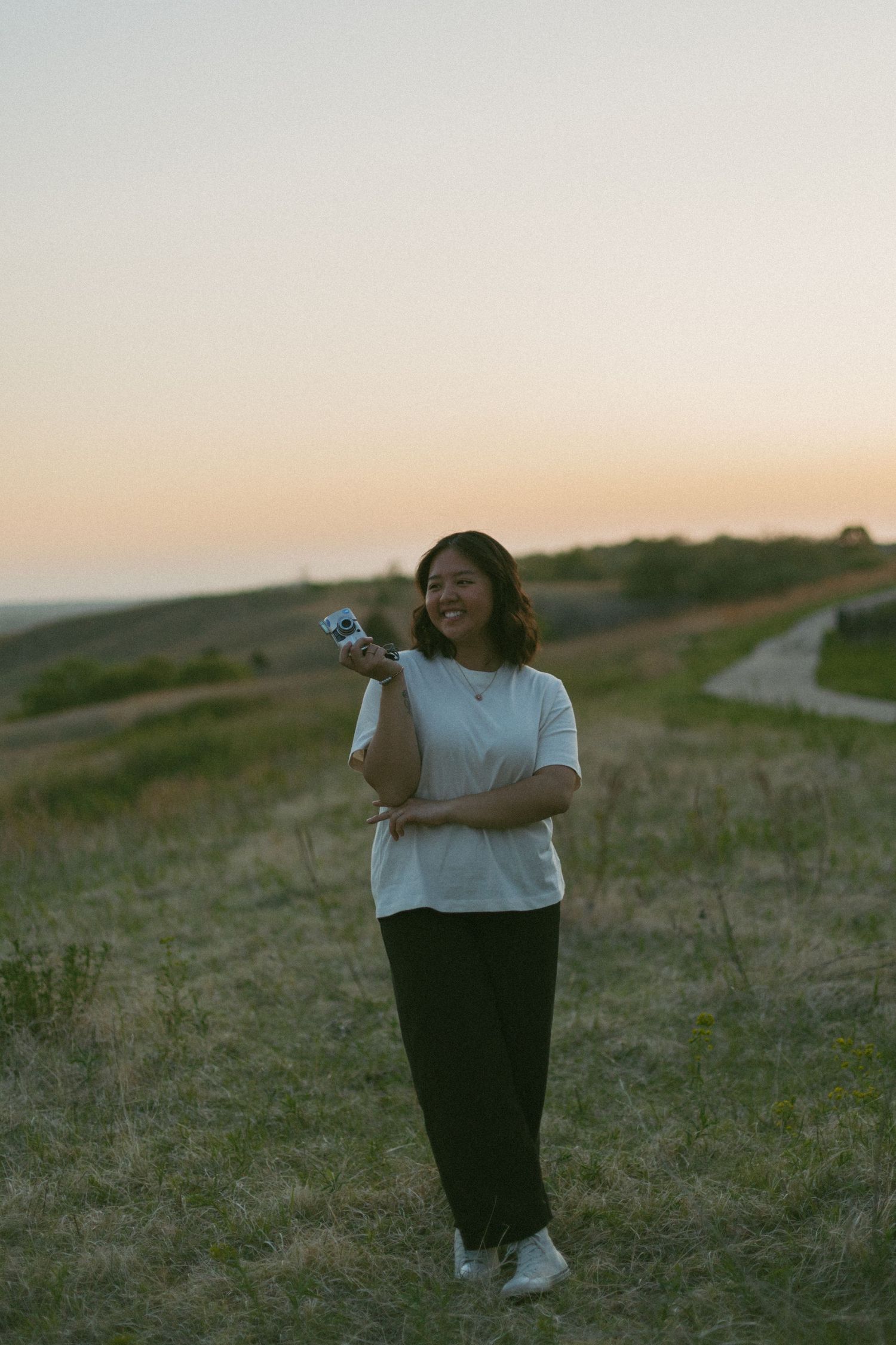 Person in a white sweater and dark pants sits on a grassy hillside during golden hour sunset.