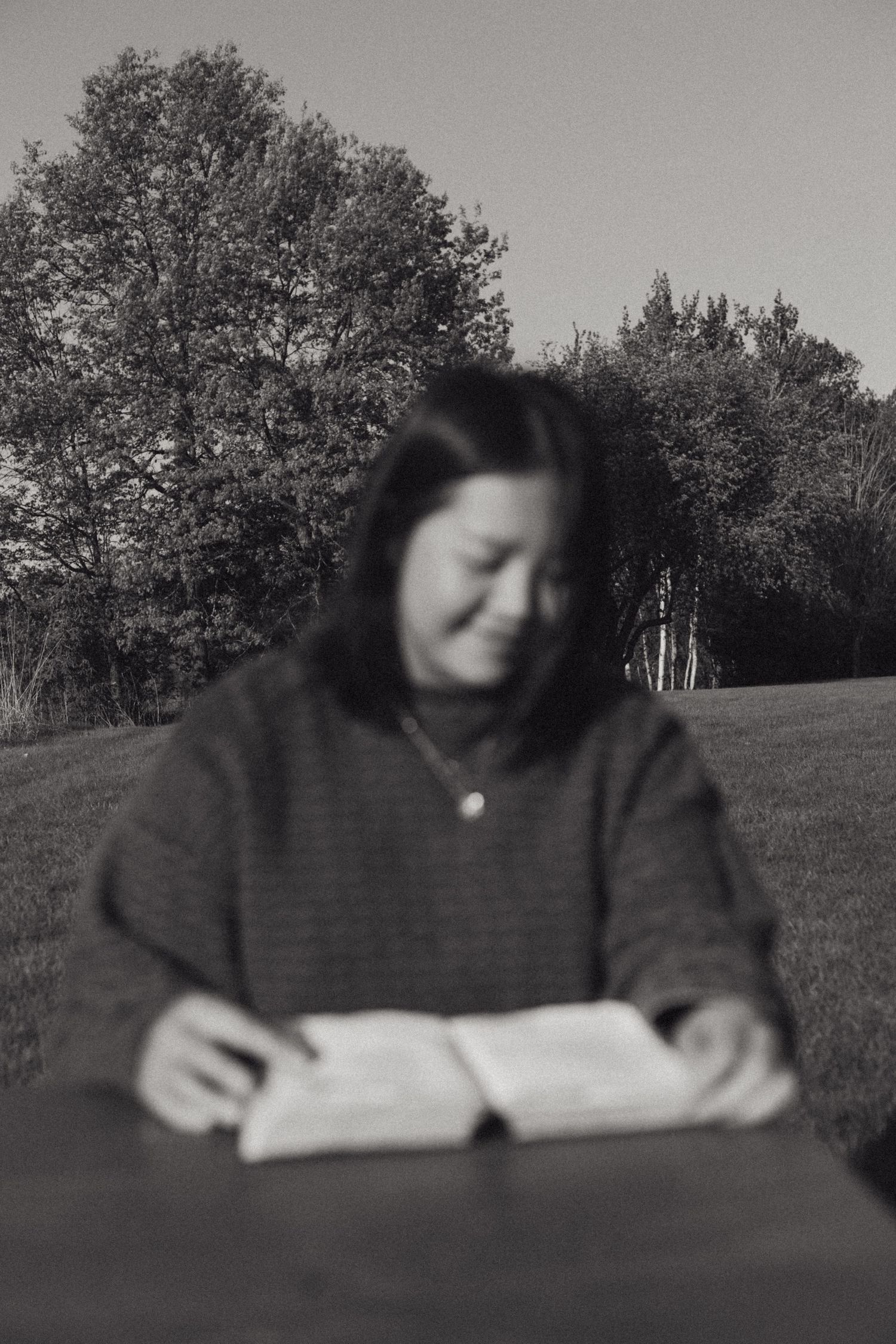 A person in dark clothing reads a book outdoors on a sunny day with trees in the background.