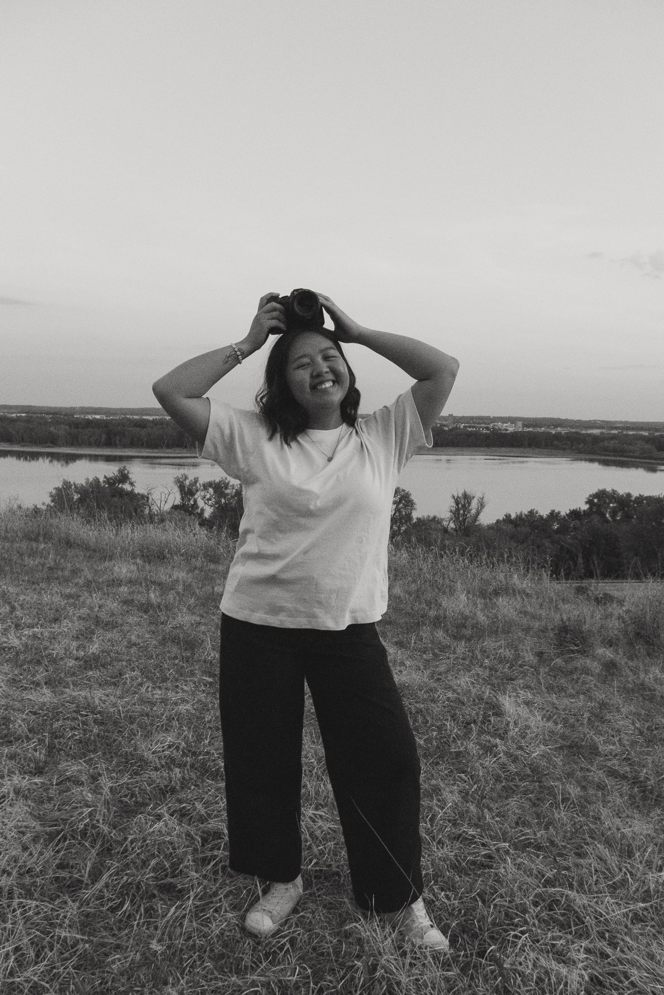 Black and white photo of someone in a white t-shirt and dark pants standing on a hilltop overlooking a lake at sunset.