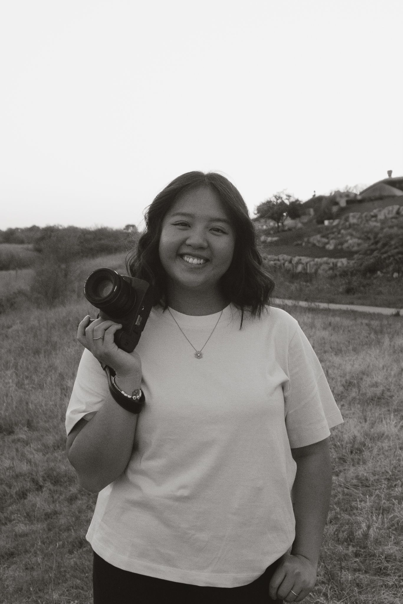 A photographer in a white t-shirt poses outdoors in a grassy field with a camera in black and white tones.