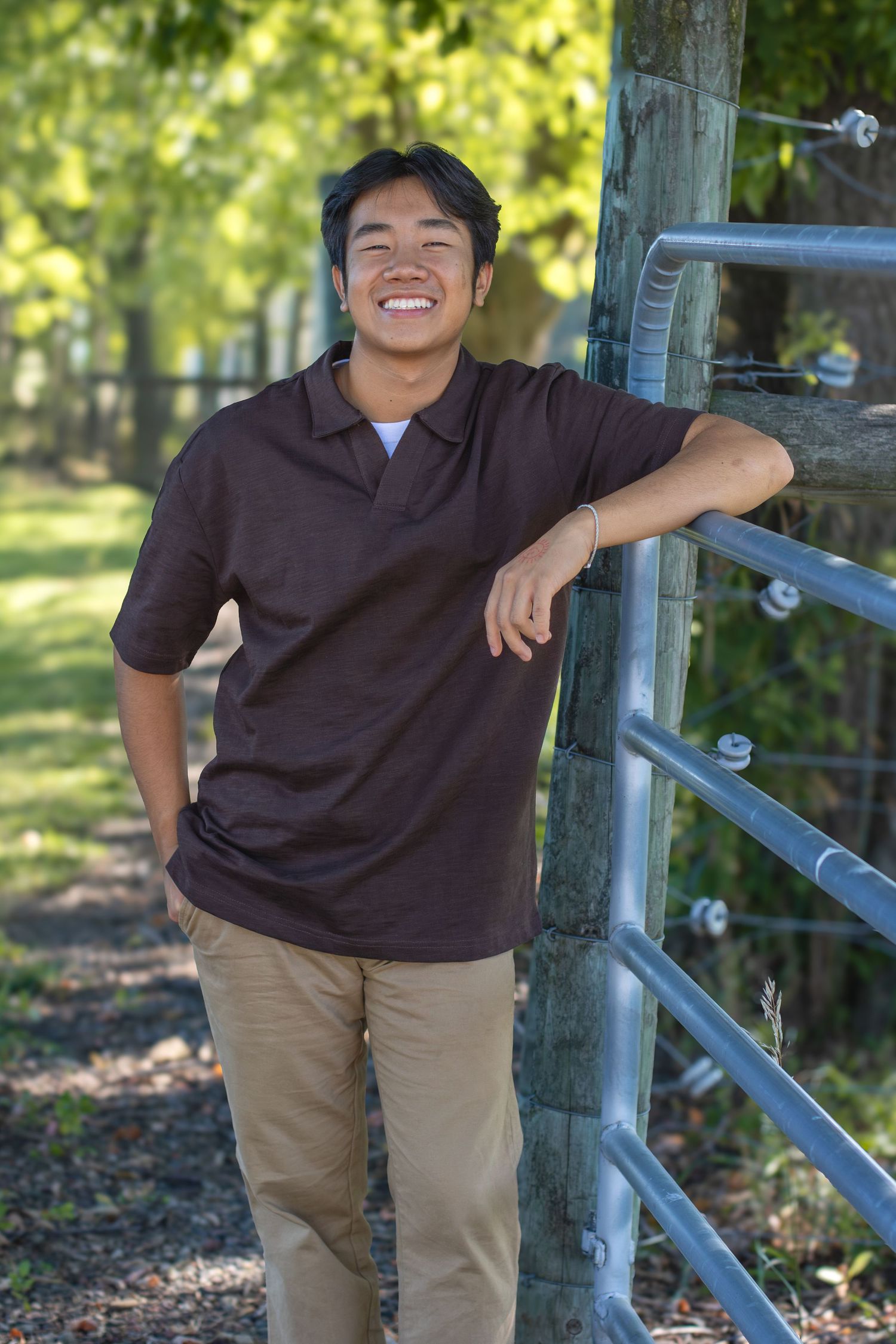 A person in a brown polo shirt and khaki pants leans against a wooden fence in a sunny outdoor setting.
