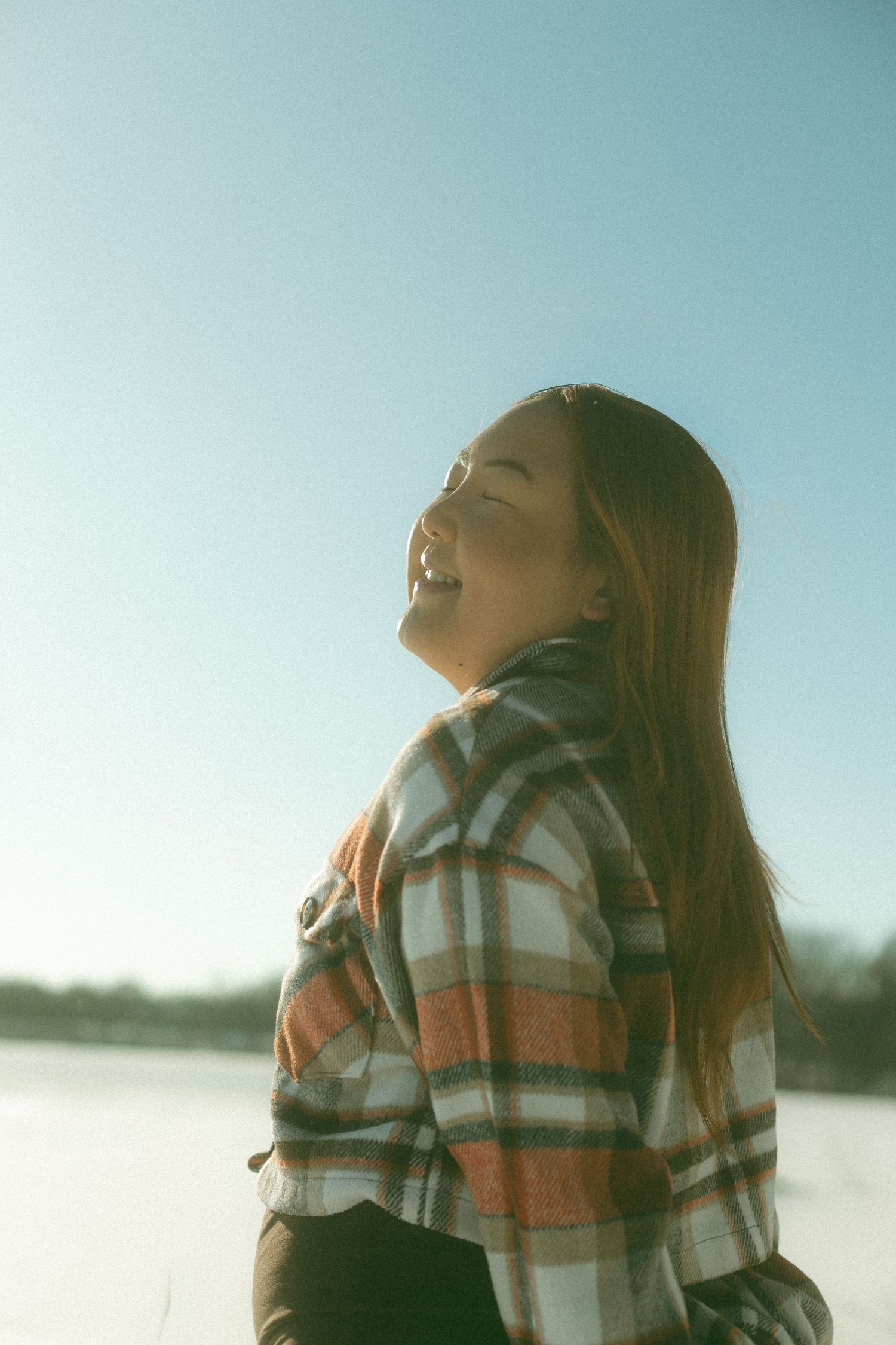 A person in a plaid shirt tilts their head back against a snowy winter landscape at sunset.