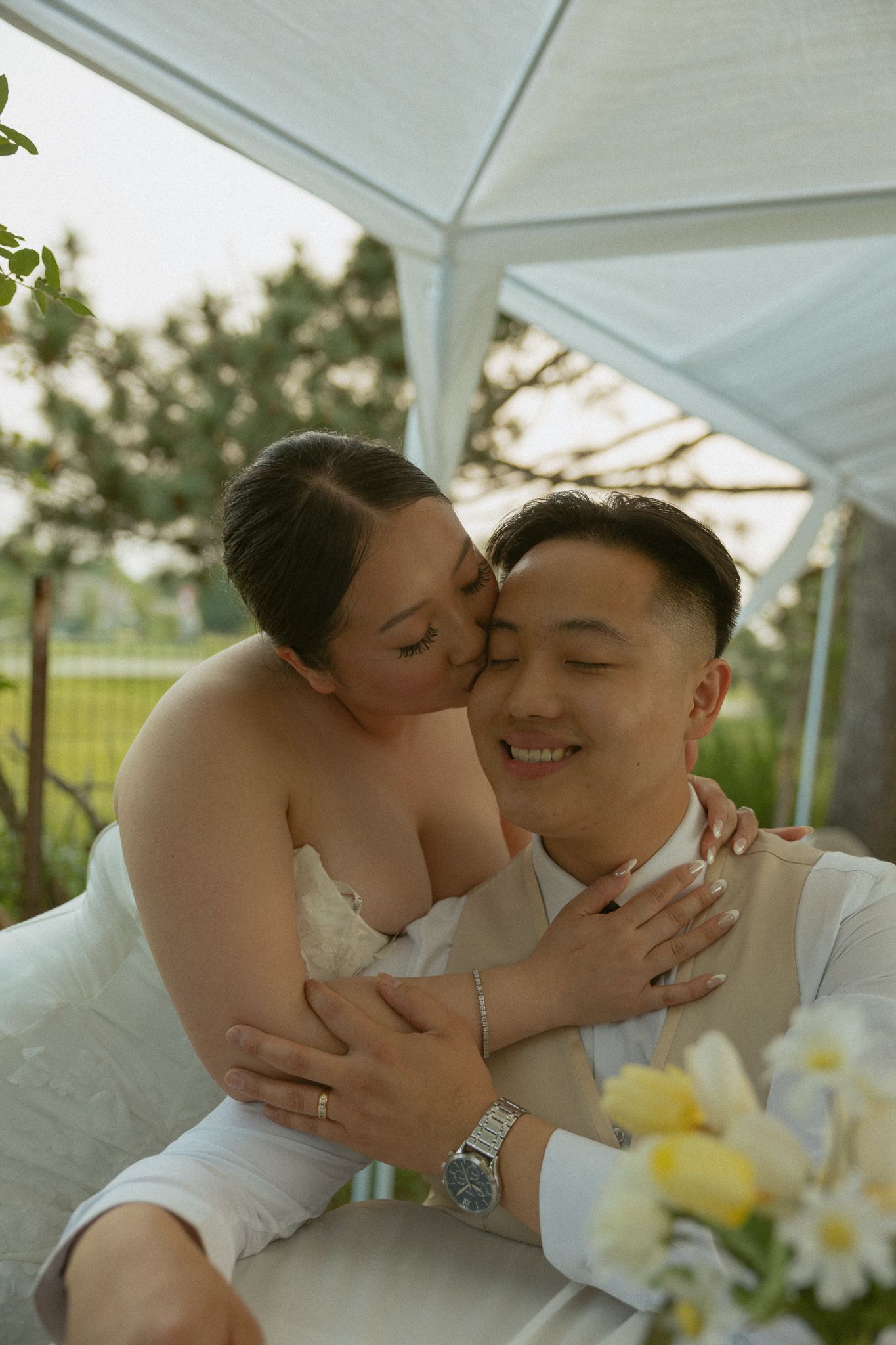 A romantic couple in white attire share tender moments at an outdoor venue decorated with yellow and white flowers.