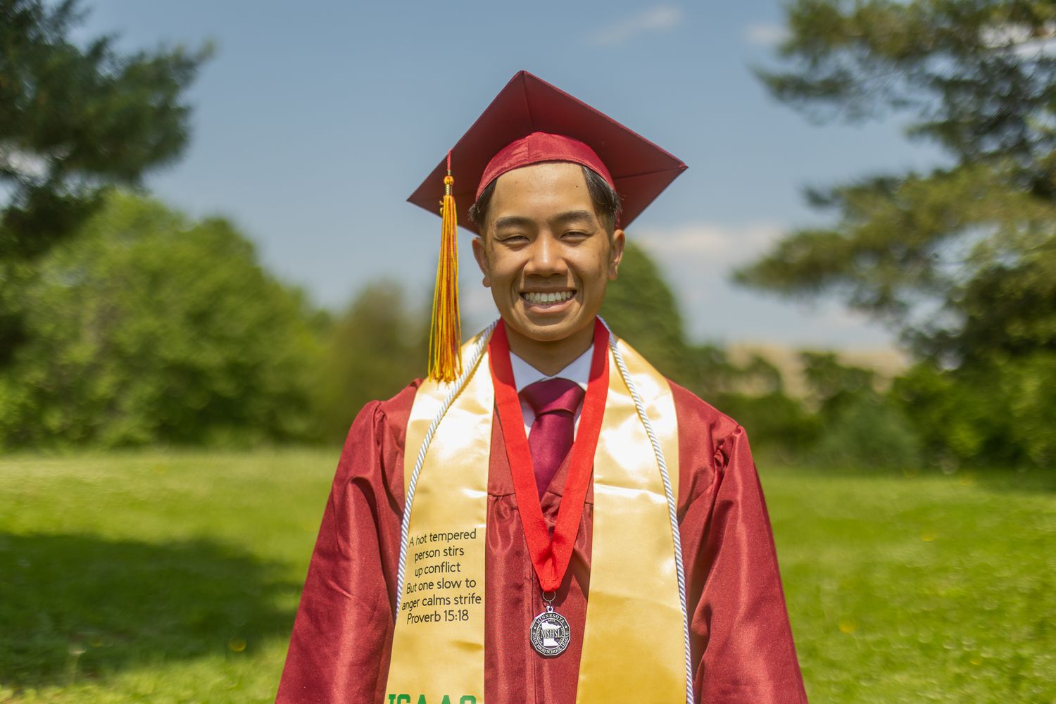 Graduate in red and gold academic regalia with honor cords and stoles celebrating outdoors.