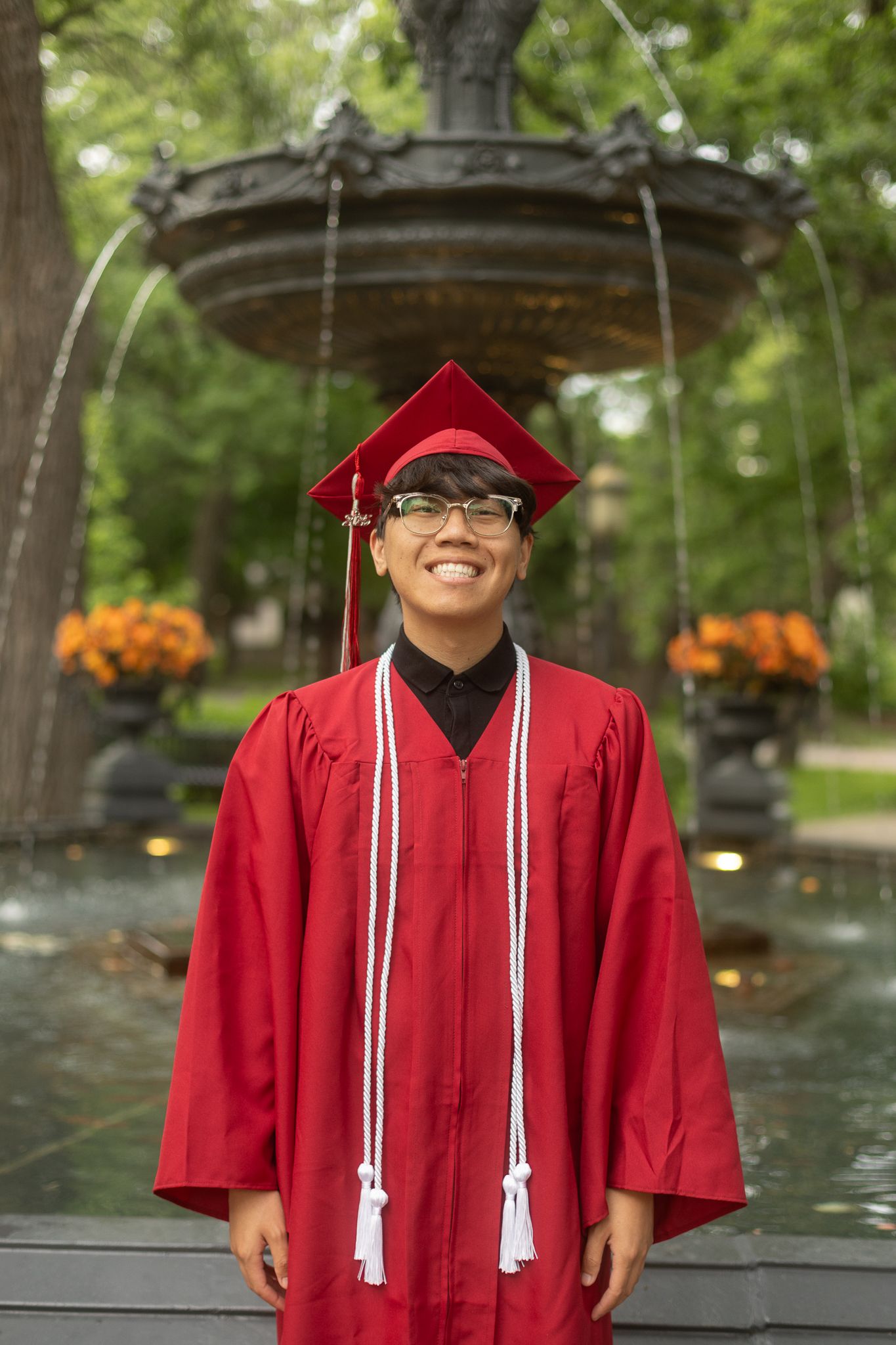 Graduate in bright red cap and gown smiling outdoors on graduation day with trees and fountains in background.