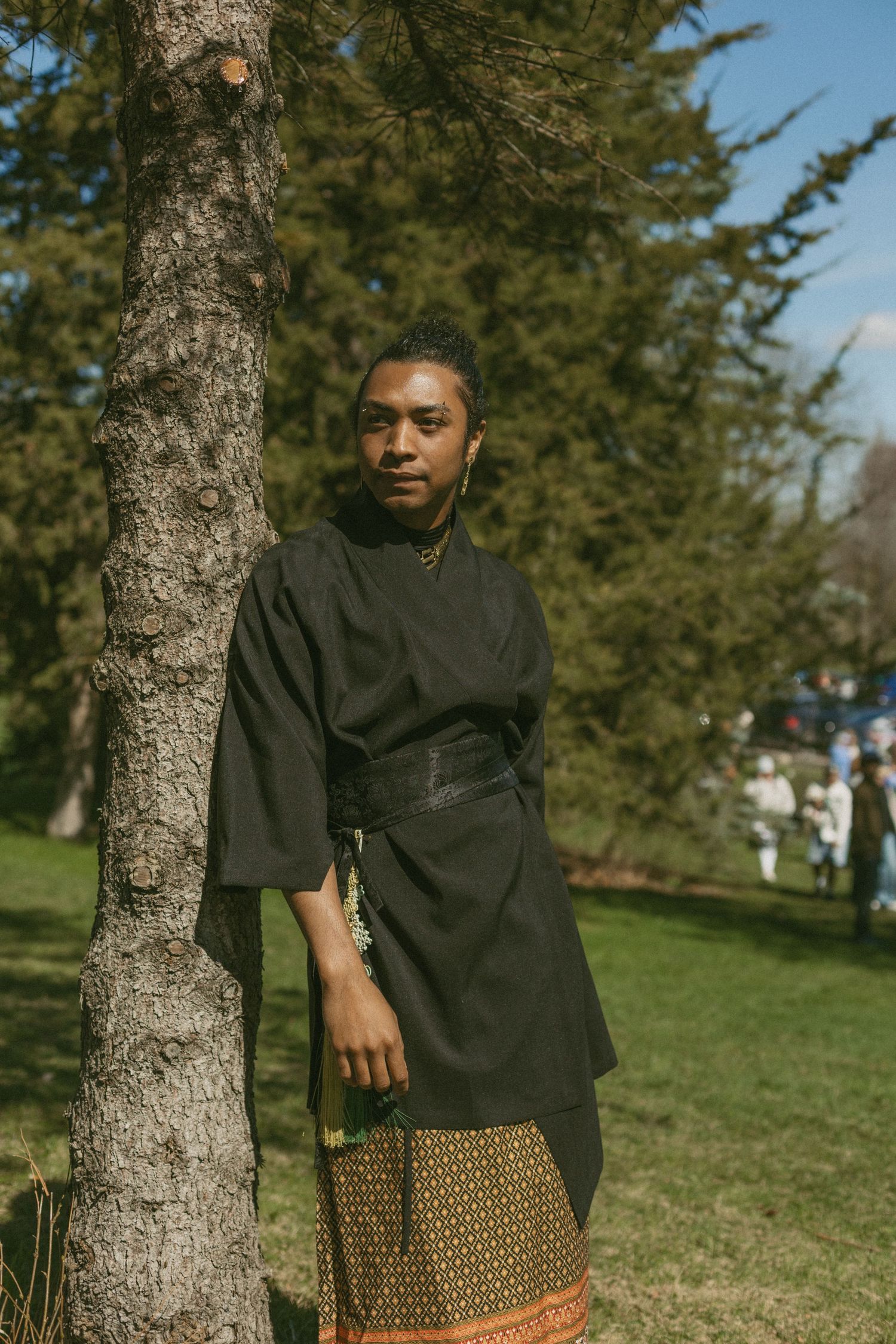 A person in a black top and patterned skirt poses thoughtfully against a tree trunk in a sunny park setting.