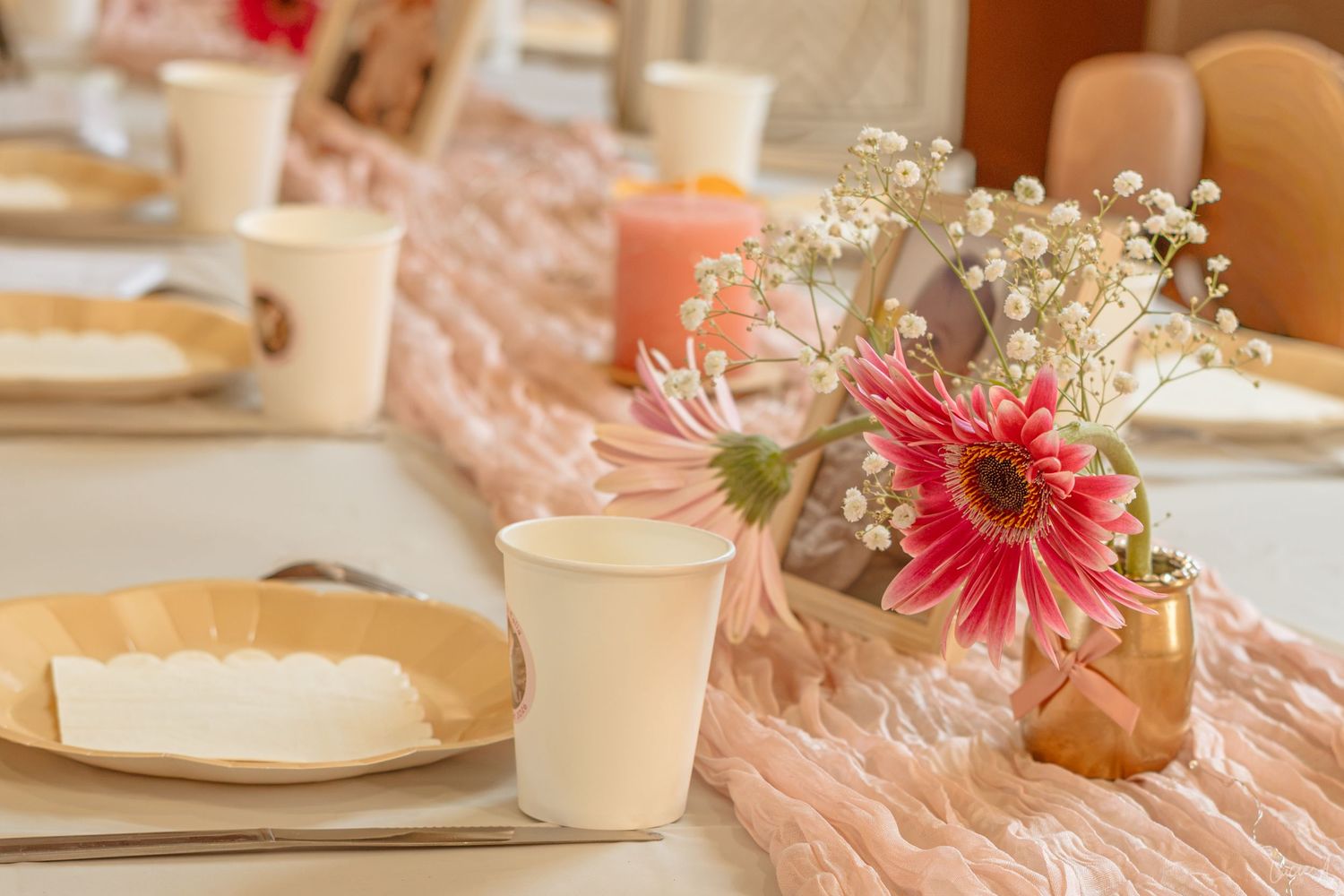 Elegant party table setting with pink gerbera daisies, baby's breath in copper vase, peach runner, paper cups and kraft plates.