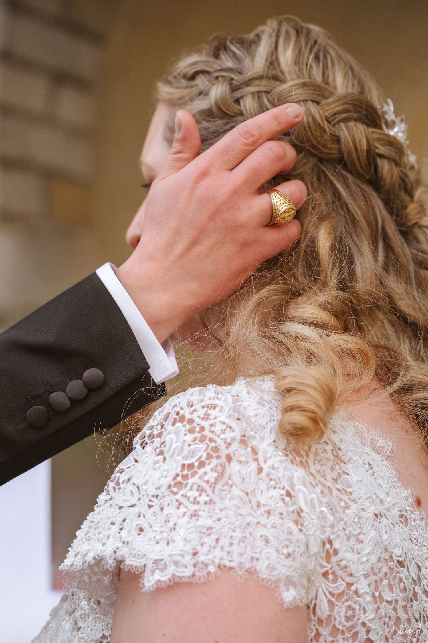 Le marié caresse tendrement la coiffure tressée de la mariée lors de la cérémonie, portant une alliance en or et une veste de costume sombre