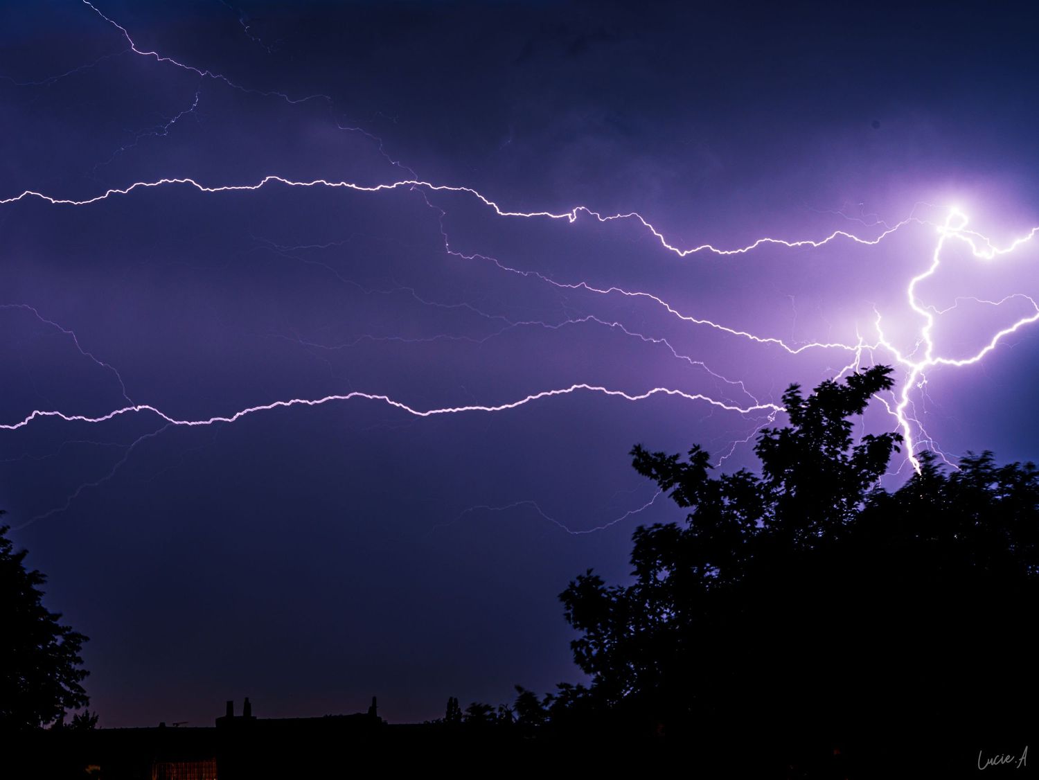 Un ciel nocturne d'un violet spectaculaire, traversé par deux éclairs horizontaux qui zèbrent les silhouettes des arbres et des toits.