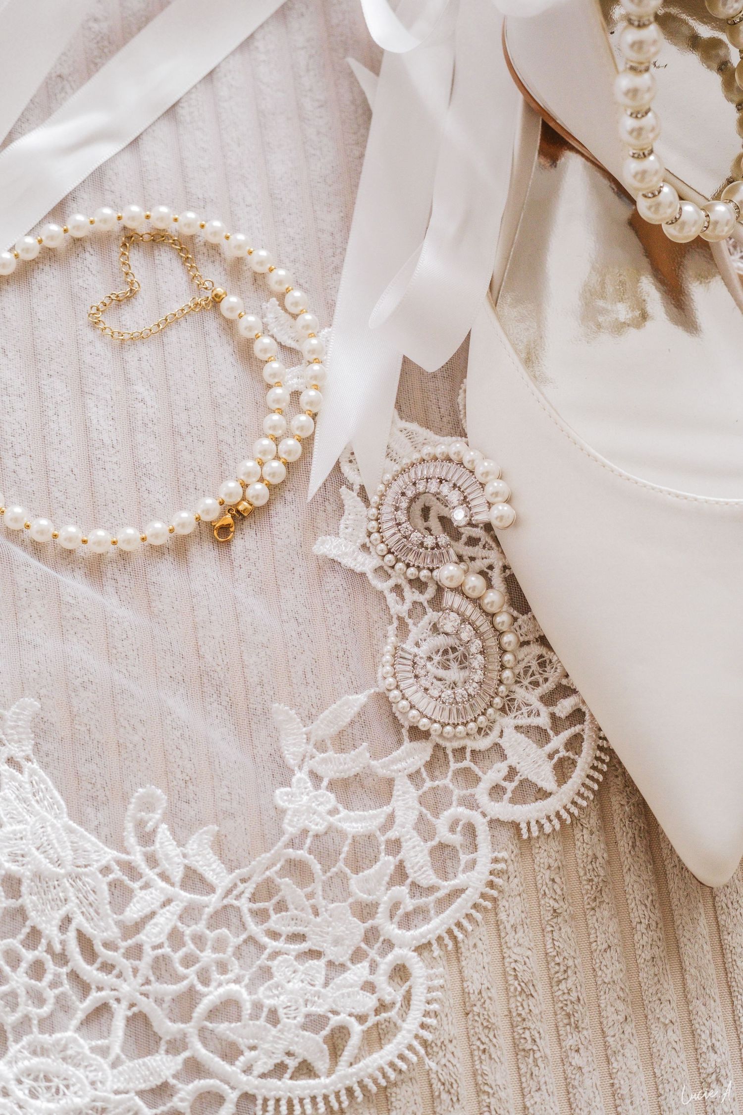 Accessoires de mariée élégants présentés à plat : bracelet de perles, voile de dentelle, talons blancs et broche en cristal sur fond ivoire.