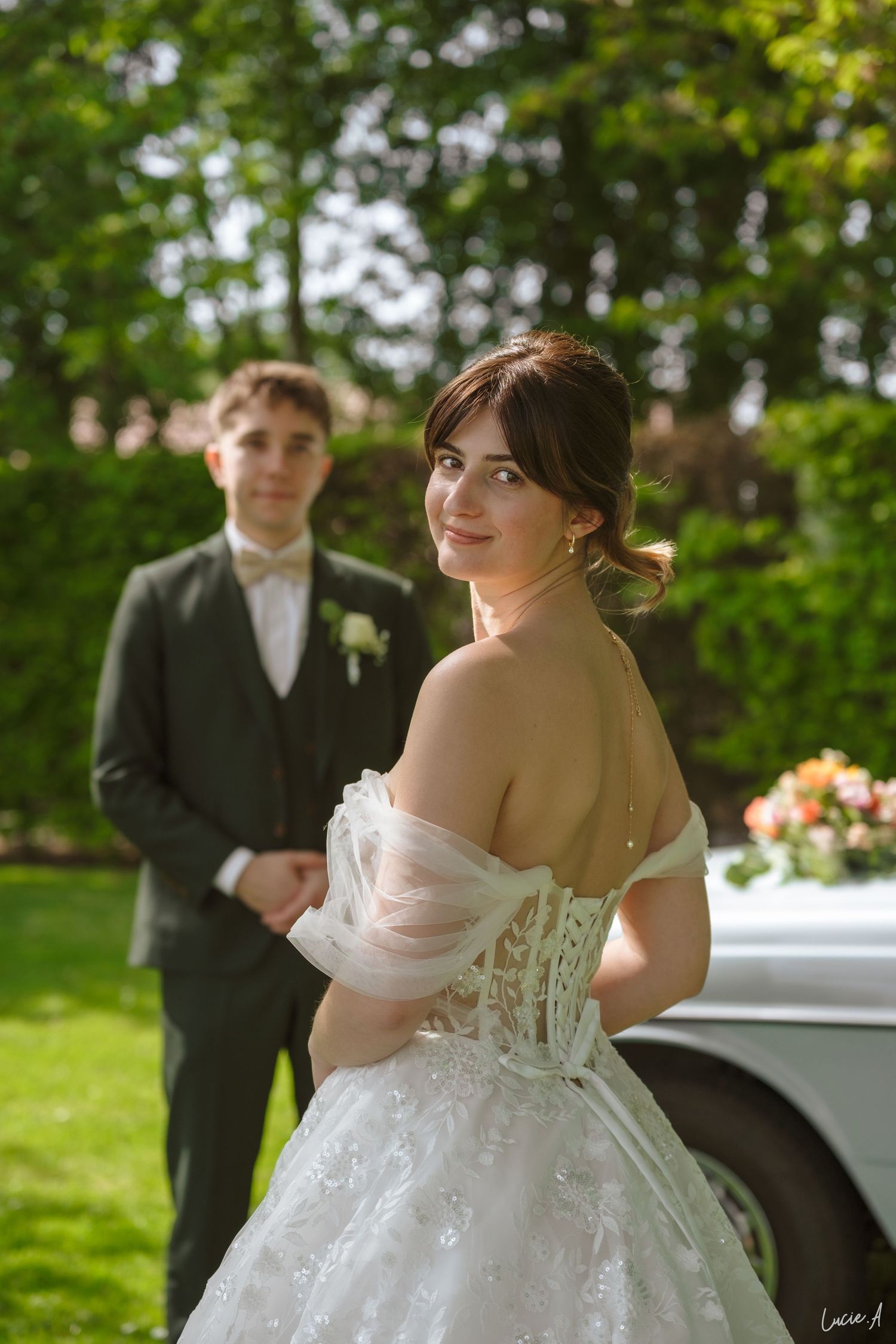 La mariée, vêtue d'une élégante robe de mariée en dentelle à épaules dénudées, sourit par-dessus son épaule, tandis que le marié, en costume