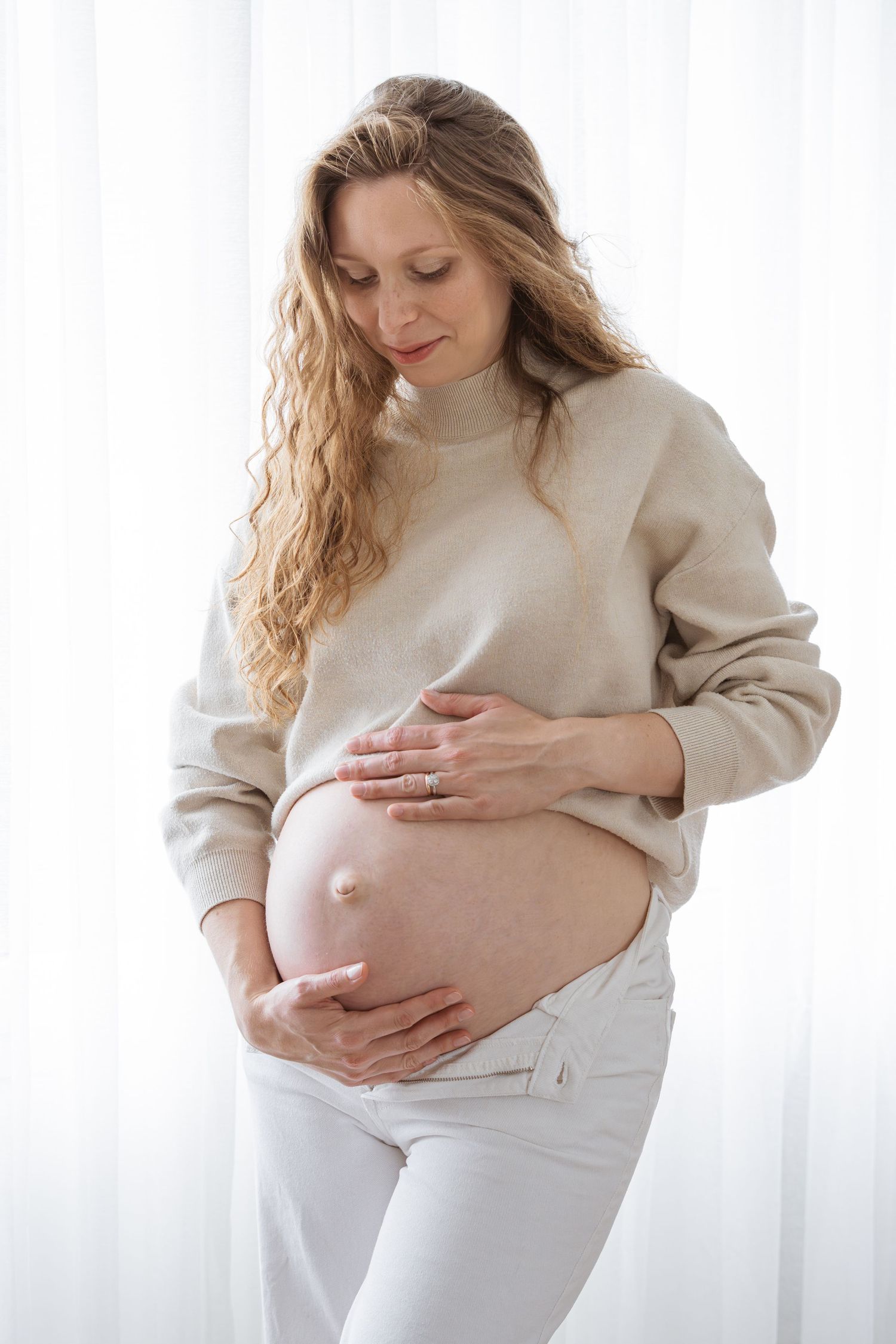 Pregnant woman in beige sweater and white pants cradling her bare baby bump with both hands near a bright window.
