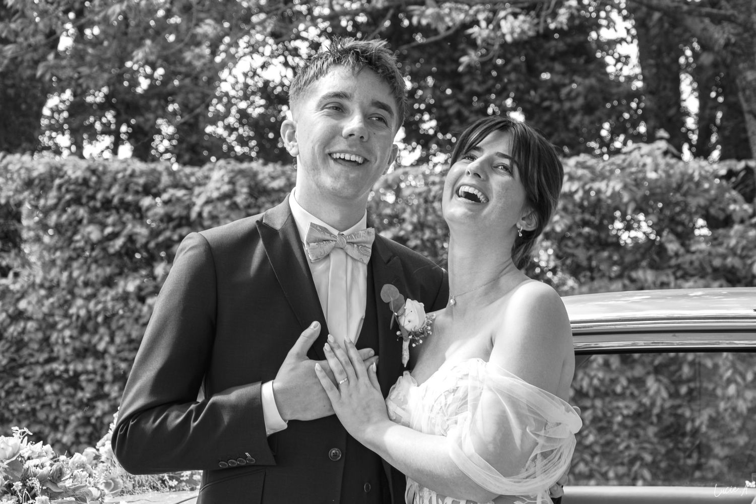 Photo en noir et blanc d'une mariée et d'un marié riant, en tenue de mariage, posant à l'extérieur près d'une végétation luxuriante.